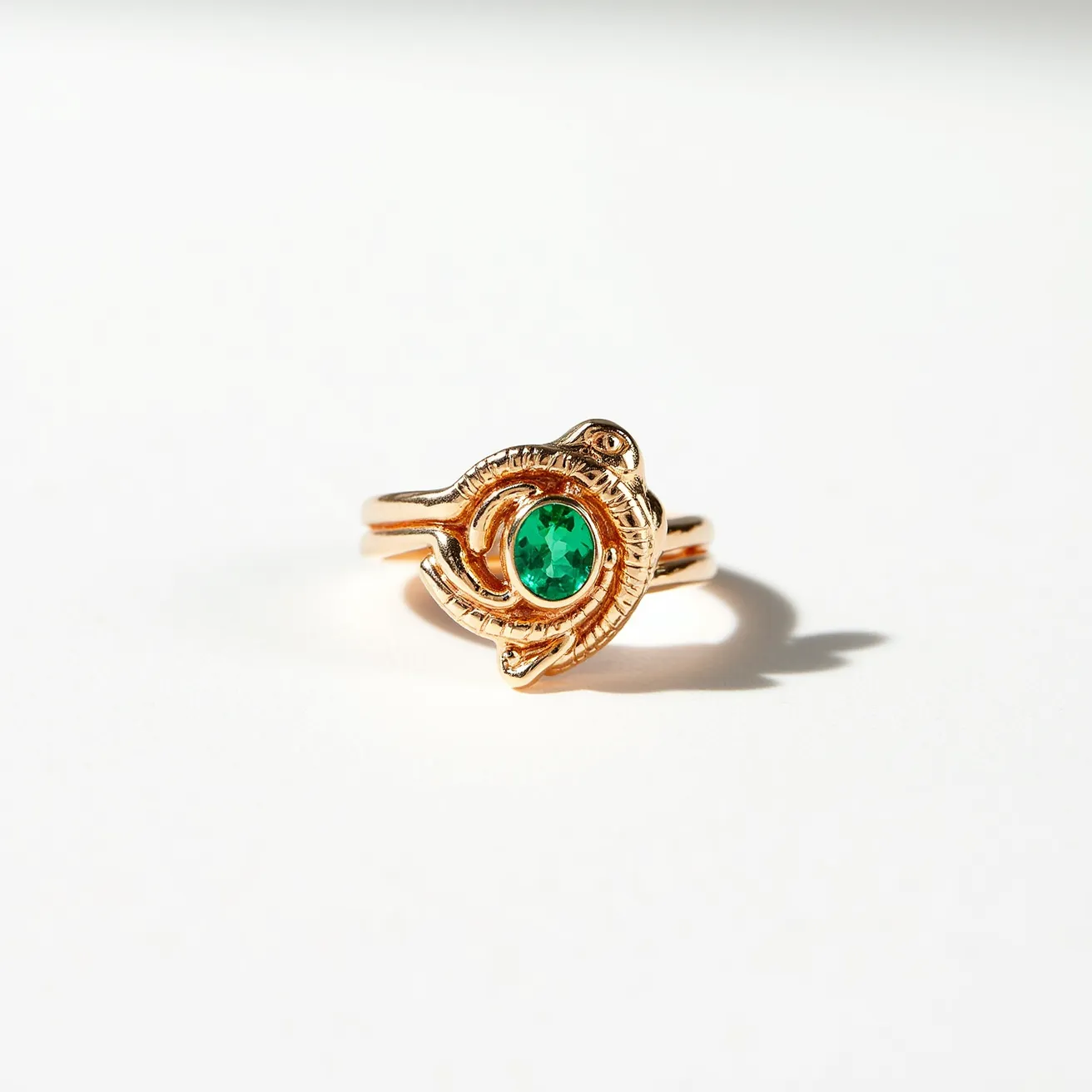 This golden bird ring showcases an intricate design where a bird motif elegantly wraps around a vibrant green gemstone, possibly an emerald. The stone is cut in an oval shape and is secured in a bezel setting, which enhances its prominent green hue. Crafted in lustrous gold, the ring's material complements the gemstone, creating a harmonious and luxurious appearance. The band is designed to seamlessly integrate the bird figure into its overall structure, providing both aesthetic appeal and a secure fit.