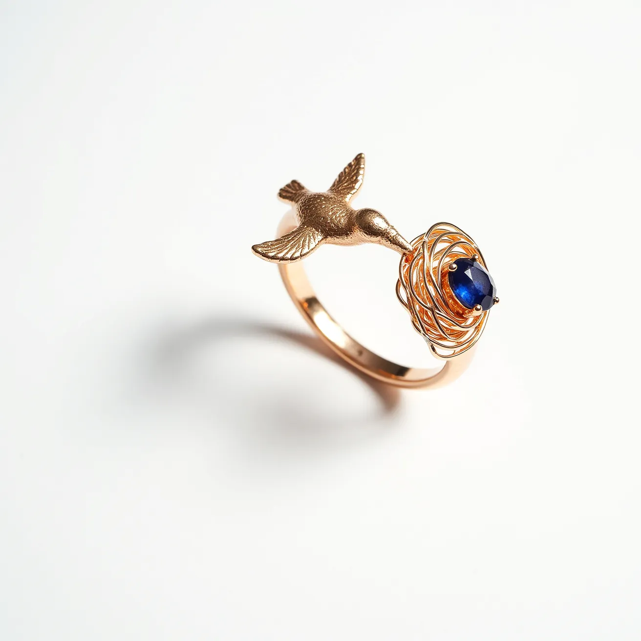 This golden bird ring elegantly showcases a detailed bird design crafted from a rich gold material. The bird appears to be flying towards a beautifully crafted nest made of intricate gold wirework. Nestled within the golden nest is a striking blue gemstone, cut in an oval shape, and set securely with prongs that elegantly hold the gem in place, adding a vibrant contrast to the overall design. The ring does not feature any visible clasps or additional attachments, highlighting its seamless and artistic form.