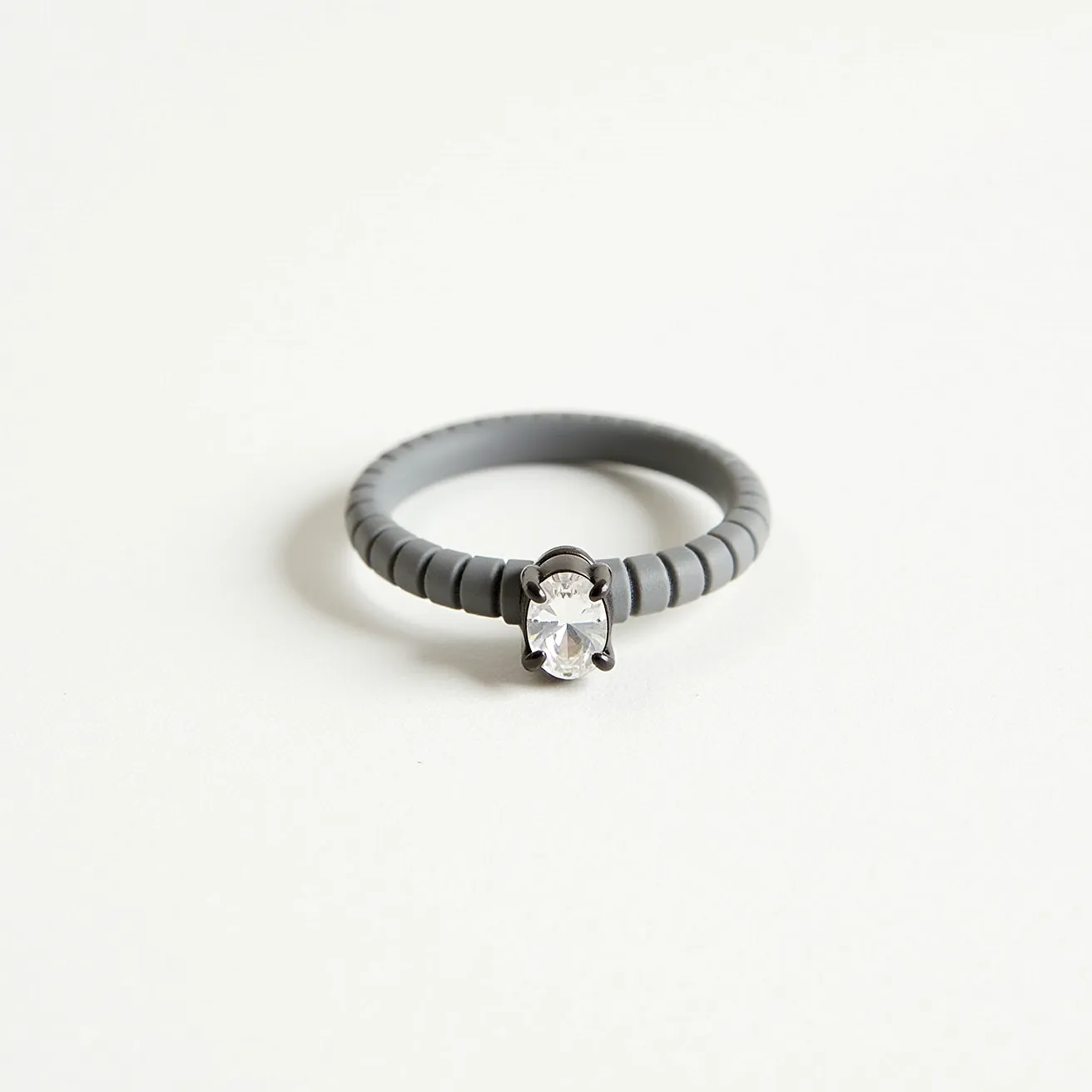 This groove ring features a sleek, textured band made of a matte, rubber-like material that adds a modern and durable touch. At the center, it showcases an oval gem, likely a clear stone, securely set in a prong setting that enhances its brilliance and catchlight. The prongs appear to be made from a contrasting, darker metal, providing a striking visual contrast against the stone and the band. This combination of materials and design creates a unique blend of style and practicality.