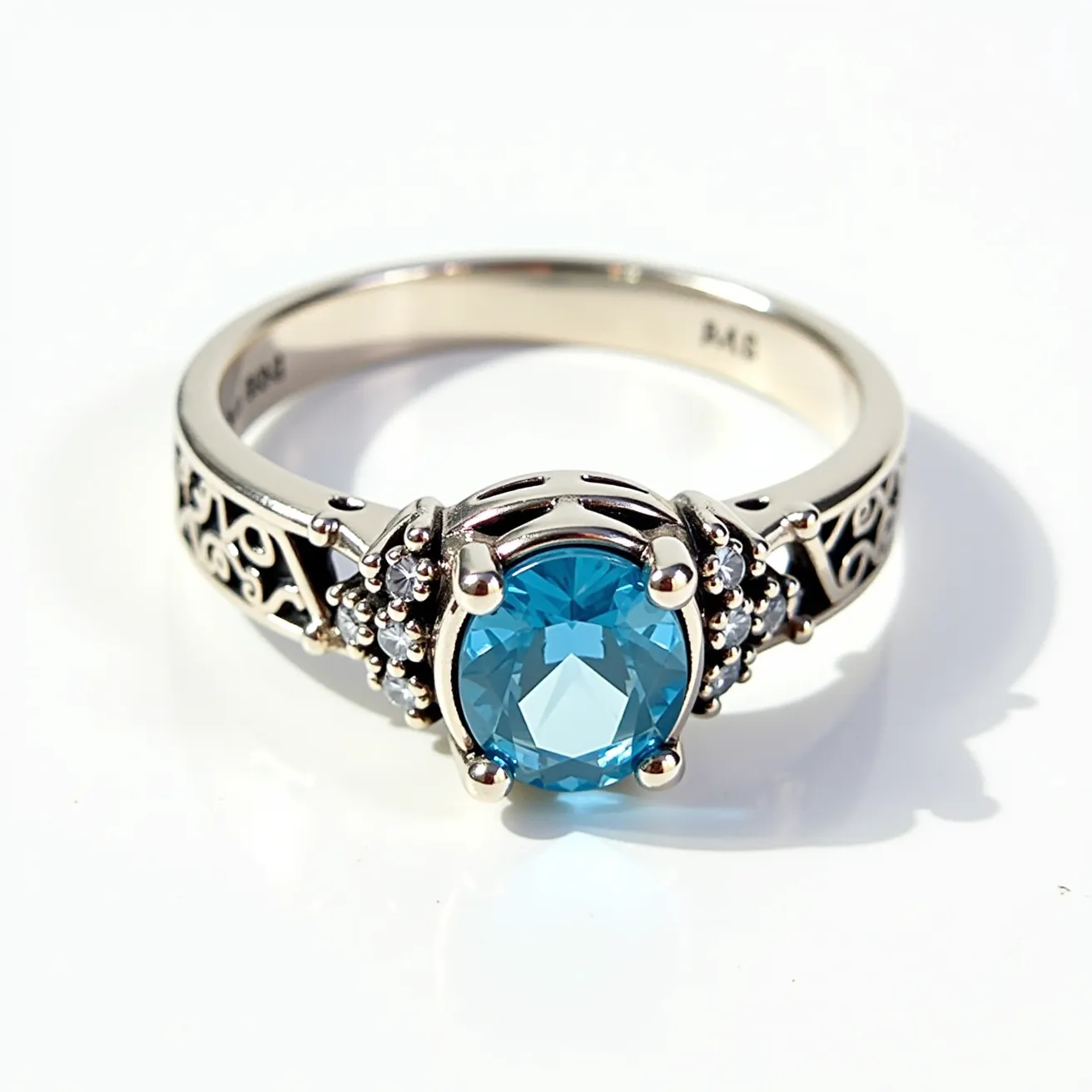 This groove ring features a prominent round-cut blue gemstone set in a prong setting, which holds the stone securely at its center. The metal appears to be a polished silver or white gold, providing a sleek and elegant framework for the ring. On either side of the main stone are intricate scrollwork patterns, adding ornate details to the band. The ring includes smaller accent stones flanking the central gem, enhancing its overall sparkle and visual interest. There is no visible clasp or attachment, as is typical with rings designed in this fashion.