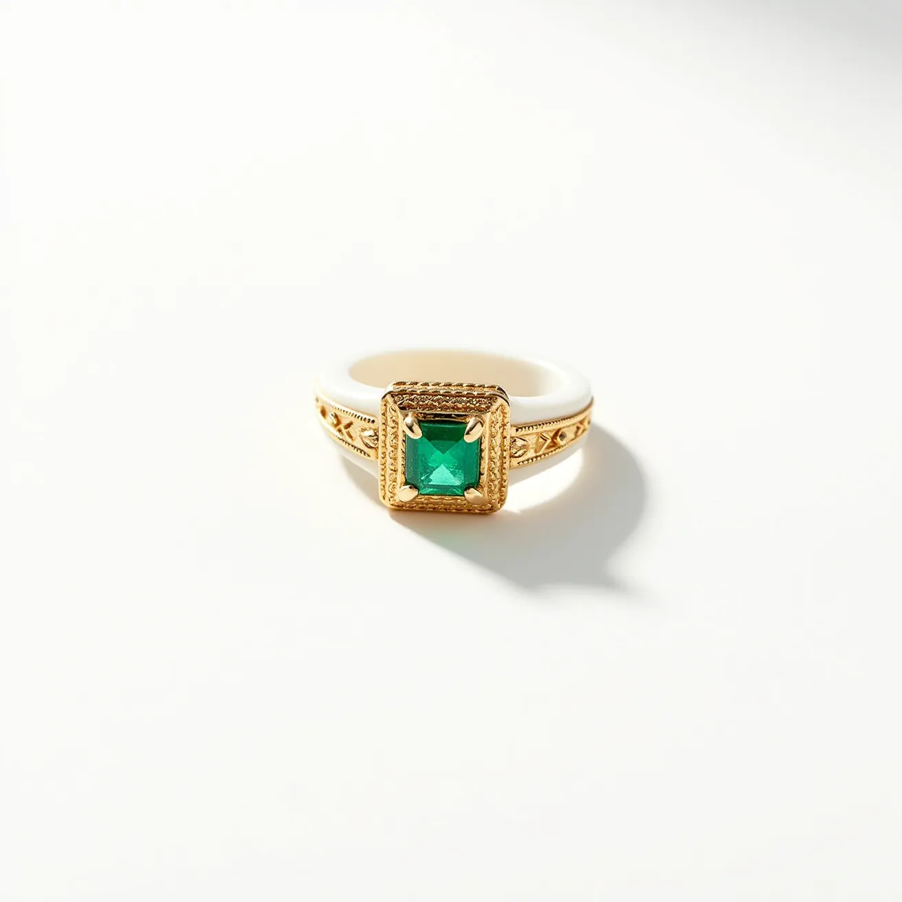 This groove ring features a striking combination of materials, with a band that appears to be made of a smooth, light-colored resin or ceramic paired with an ornate gold-toned metal setting. The centerpiece of the ring is a vividly colored square-cut green gemstone, likely an emerald, which is set in a square bezel showcasing a pattern of intricate detailing around the stone. The gold-toned setting incorporates decorative elements that add to the elegance, while the main gemstone is firmly held by several prongs, ensuring stability. The overall design of the ring merges classic elegance with modern flair, making it a standout accessory.