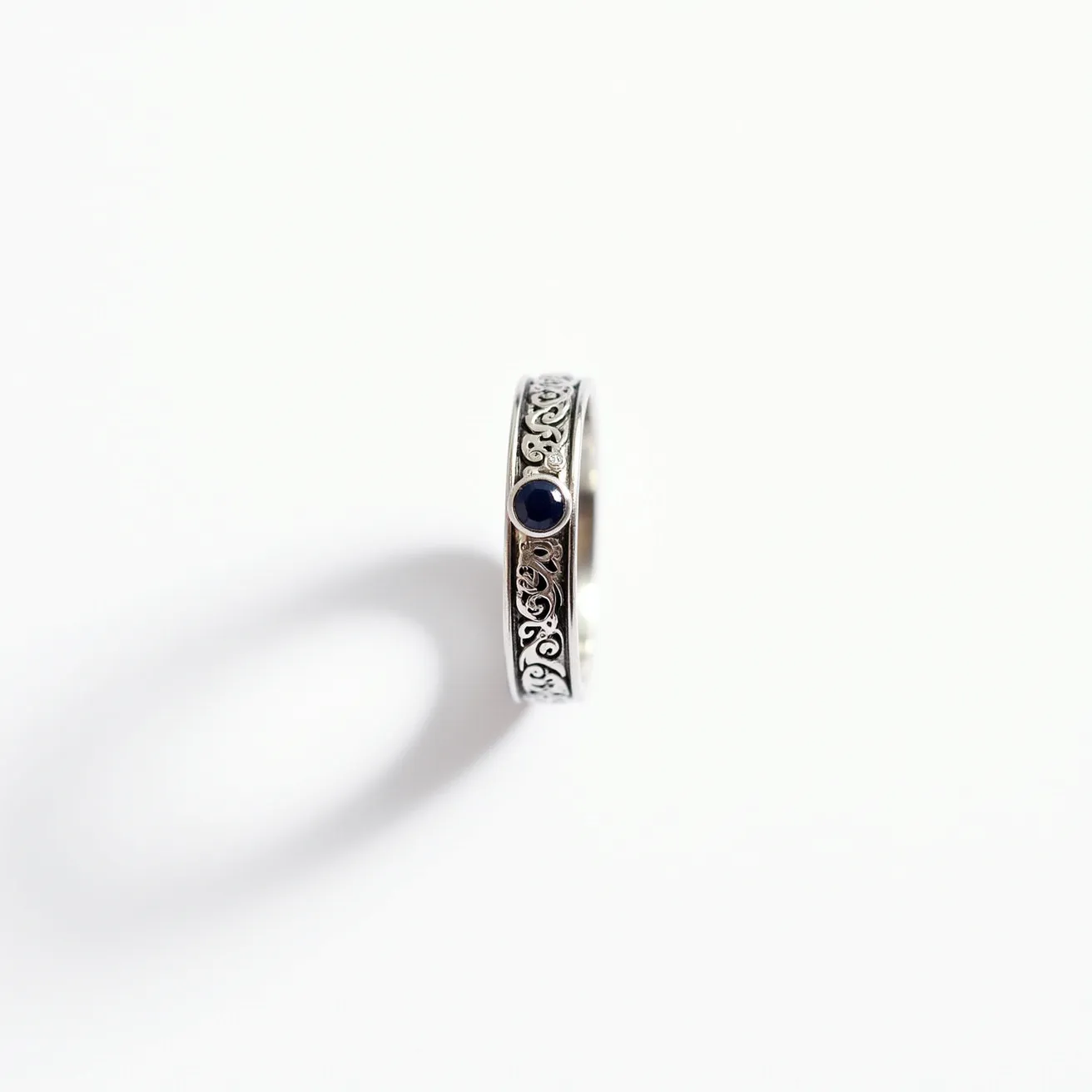 This groove ring is crafted from what appears to be a silver-toned metal, featuring an intricate filigree design along its band that adds an elegant touch. At the center of the band, there is a dark blue stone, likely a sapphire, which is round cut and bezel set, providing a secure and sleek appearance. The combination of the ornate metalwork and the deep blue gemstone creates a striking and sophisticated aesthetic, making it suitable for both formal and everyday wear.