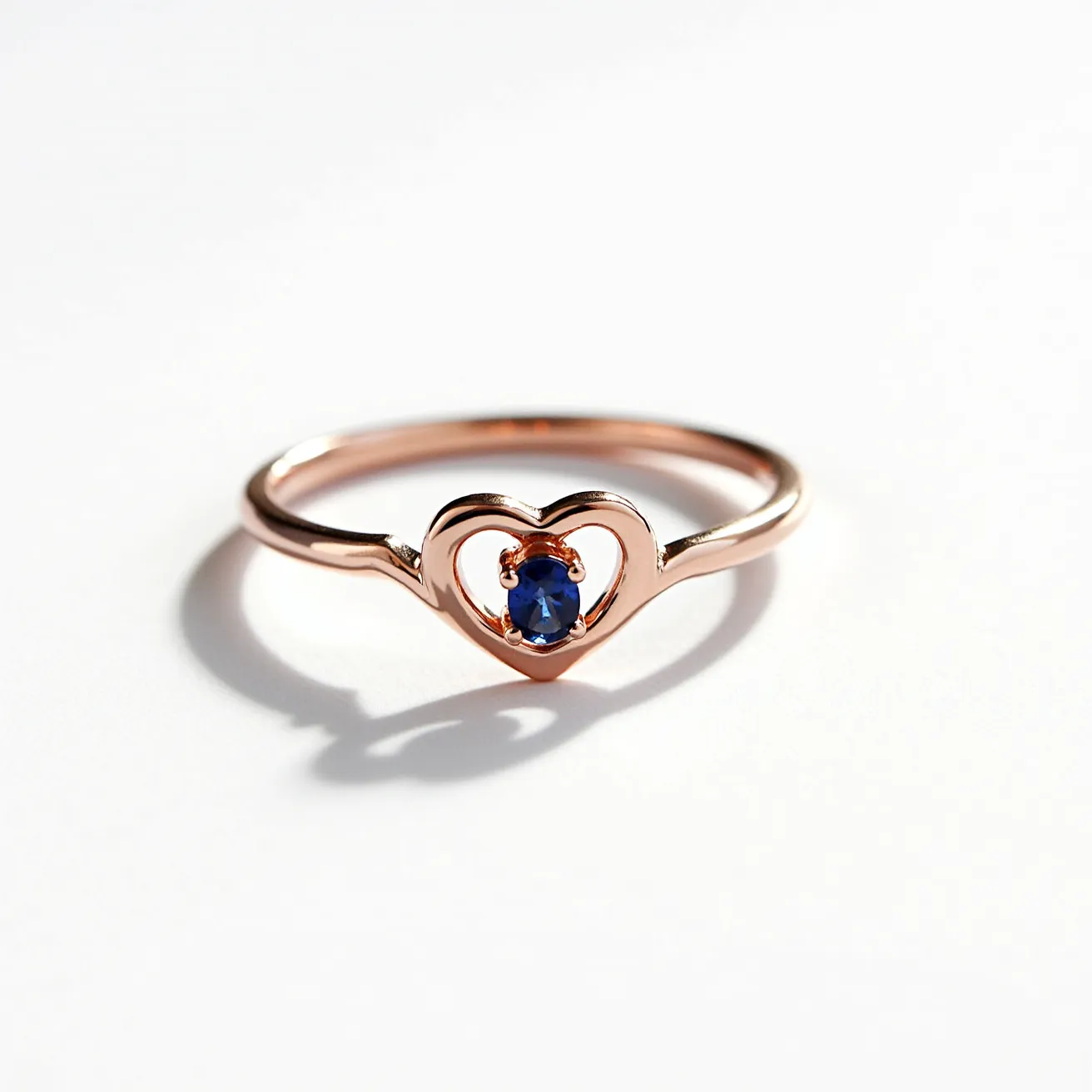 This heart promise ring is crafted from a warm rose gold metal, forming a delicate band that features a distinctive open heart design at its center. In the center of the heart rests a striking round blue gemstone, likely a sapphire, held securely in place by a classic four-prong setting. The simplicity of the ring’s design emphasizes the brilliance of the gemstone while the warm tones of the rose gold complement the deep blue of the stone, creating a harmonious and elegant piece.