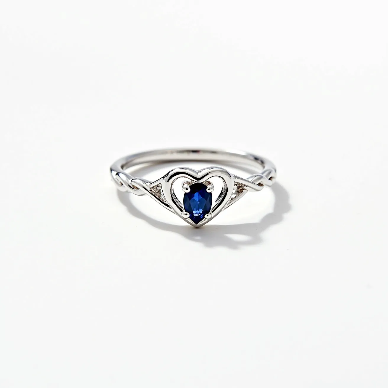 This heart promise ring features a sleek and polished band crafted from what appears to be sterling silver or another high-quality white metal. At its center is an elegantly set heart-shaped design, holding an oval blue gemstone, likely a sapphire, fixed in place by a four-prong setting. The band is complemented by a subtle twist detail, adding a touch of intricacy to its design. The overall composition of the ring highlights the central gem, making it a distinctive piece that symbolizes commitment.