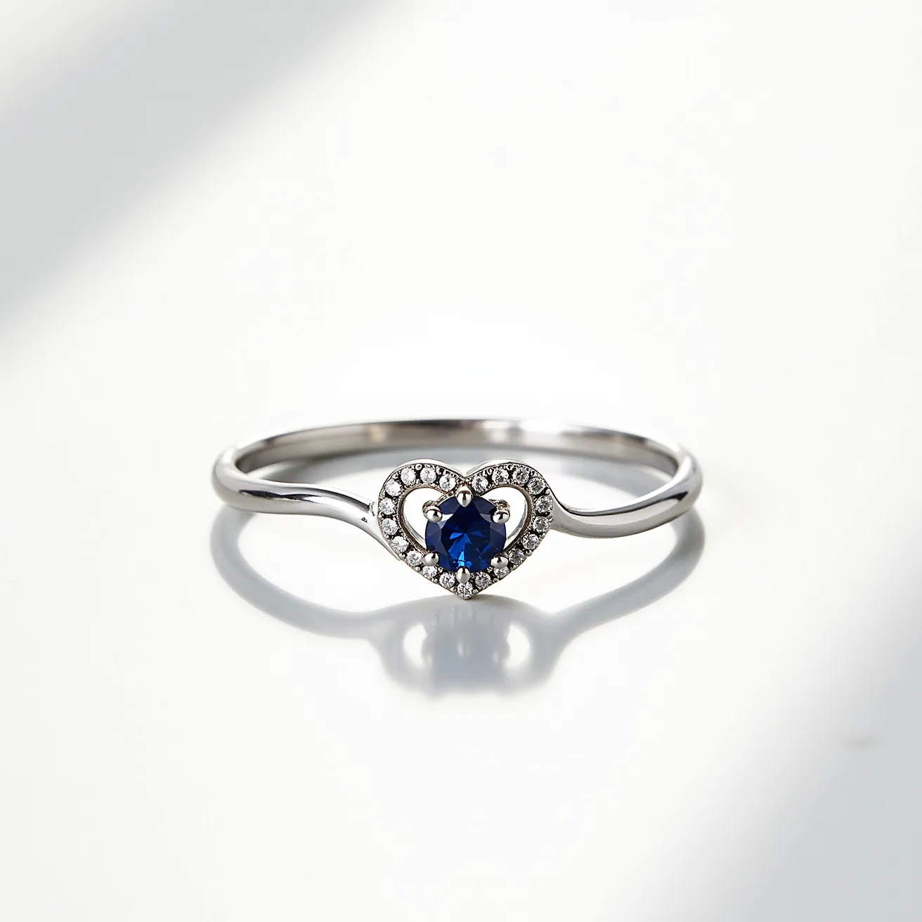 This heart promise ring features a sleek metal band, likely in a polished silver or white gold material, accentuating its elegant design. At its center, the ring showcases a beautifully cut blue gemstone, possibly a sapphire, set in a prong setting that maximizes its luminosity and depth. Surrounding the central stone is a delicate halo of small, sparkling clear gems, likely diamonds or high-quality cubic zirconia, arranged in a heart-shaped setting that enhances the romantic symbolism of the piece. The ring is designed without any visible clasps or attachments, emphasizing its minimalist and seamless aesthetic.