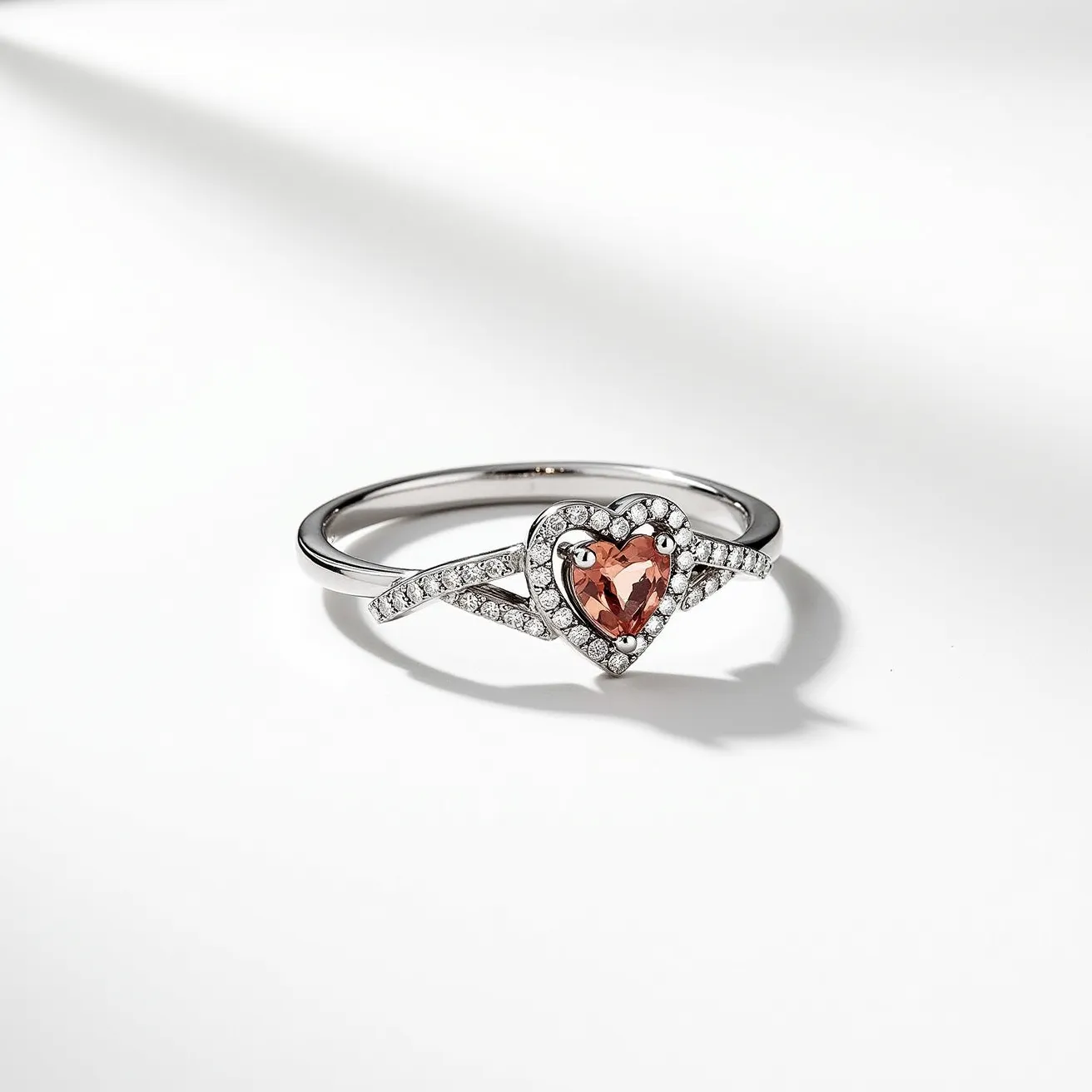 This heart ring features a central heart-shaped gem with a peach or pink hue, possibly a morganite, held in a prong setting. Surrounding the central stone is an elegant halo of smaller clear stones, likely diamonds, intricately set, enhancing the brilliance of the centerpiece. The band, likely crafted from white gold or platinum, splits gracefully as it approaches the heart, adding a delicate yet sophisticated design element. Each side of the split shank is also adorned with a row of clear stones that complement the halo, creating a cohesive, sparkling effect across the ring. The blend of the colorful heart stone with the shimmering clear stones and high-polished metal gives this ring a romantic and luxurious touch.