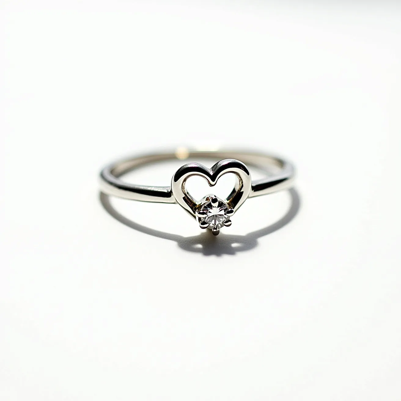 This heart ring is elegantly crafted from a smooth silver metal band, forming an open heart shape as its central feature. Nestled within the heart is a sparkling round-cut gemstone, likely a diamond, set in a minimalist setting that allows maximum light to enhance its brilliance. The stone's placement is secure, ensuring it is both visually striking and well-protected. The overall design is sleek, with a polished finish that highlights the contrast between the metal and the gemstone's shimmer, making it a timeless and graceful piece.