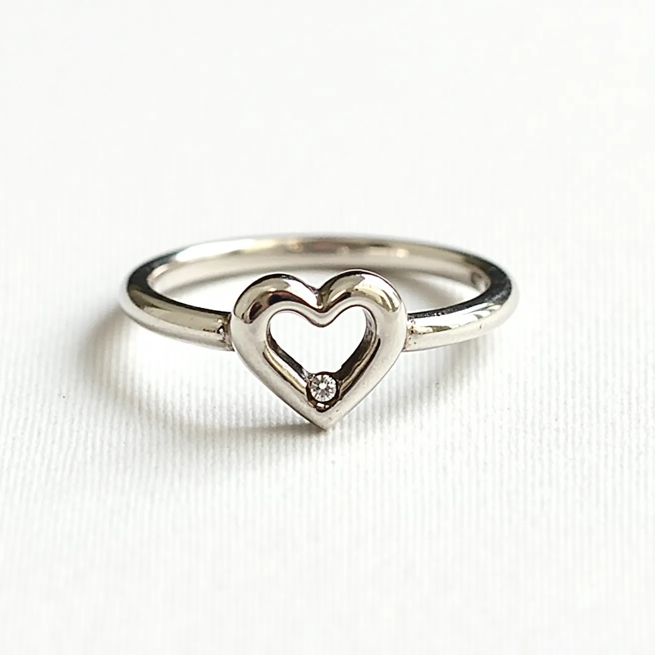 This heart ring features a sleek, polished band likely made of white gold or platinum, which enhances its elegant appearance. The centerpiece is a heart-shaped outline that is beautifully crafted and serves as a minimalistic yet striking motif. Within the open heart lies a small, round-cut diamond, set in a bezel setting that adds a touch of sparkle to the design. The ring has a seamless design without any additional clasps or attachments, emphasizing simplicity and sophistication.