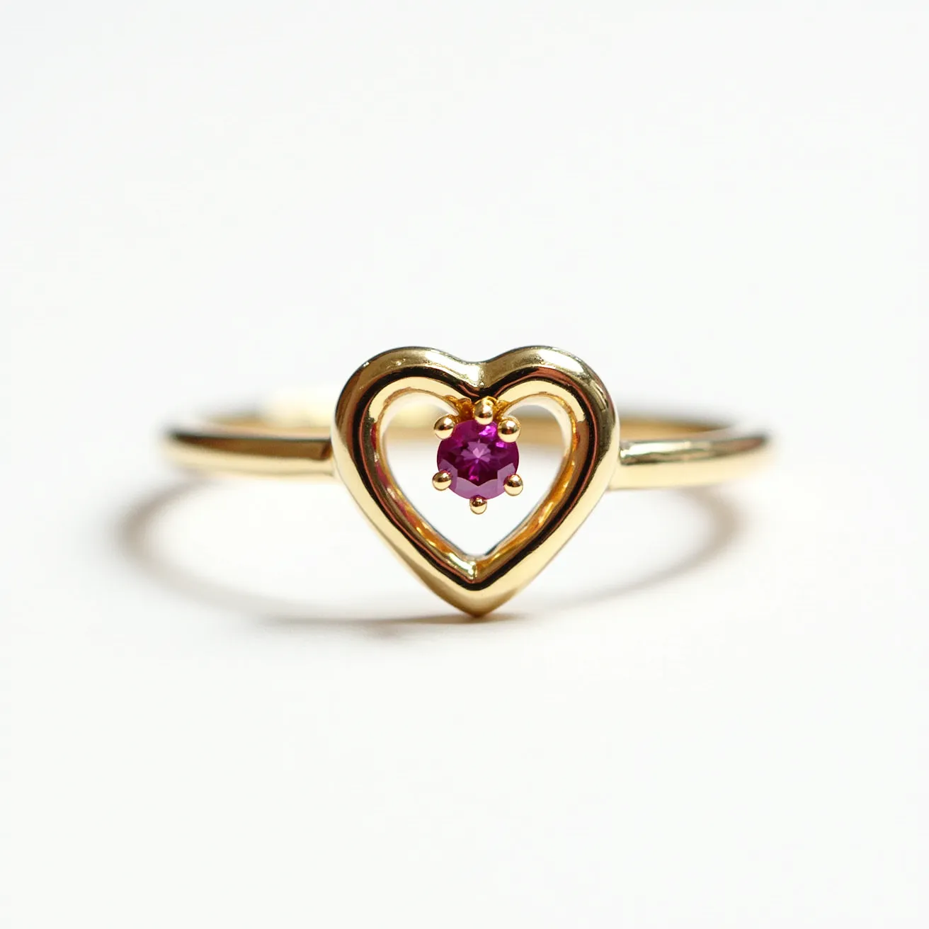 This heart ring is crafted from gold, featuring a dainty and elegant heart-shaped design. At the center of the heart, a vibrant pink gemstone, likely a ruby or pink sapphire, is prominently displayed in a round cut. The gemstone is securely set with prongs, enhancing its brilliance and ensuring its stability. The ring's band is slender and complements the minimalist yet sophisticated aesthetic of the heart and central stone arrangement. The open heart design adds a touch of modern elegance to the classic gemstone setting.