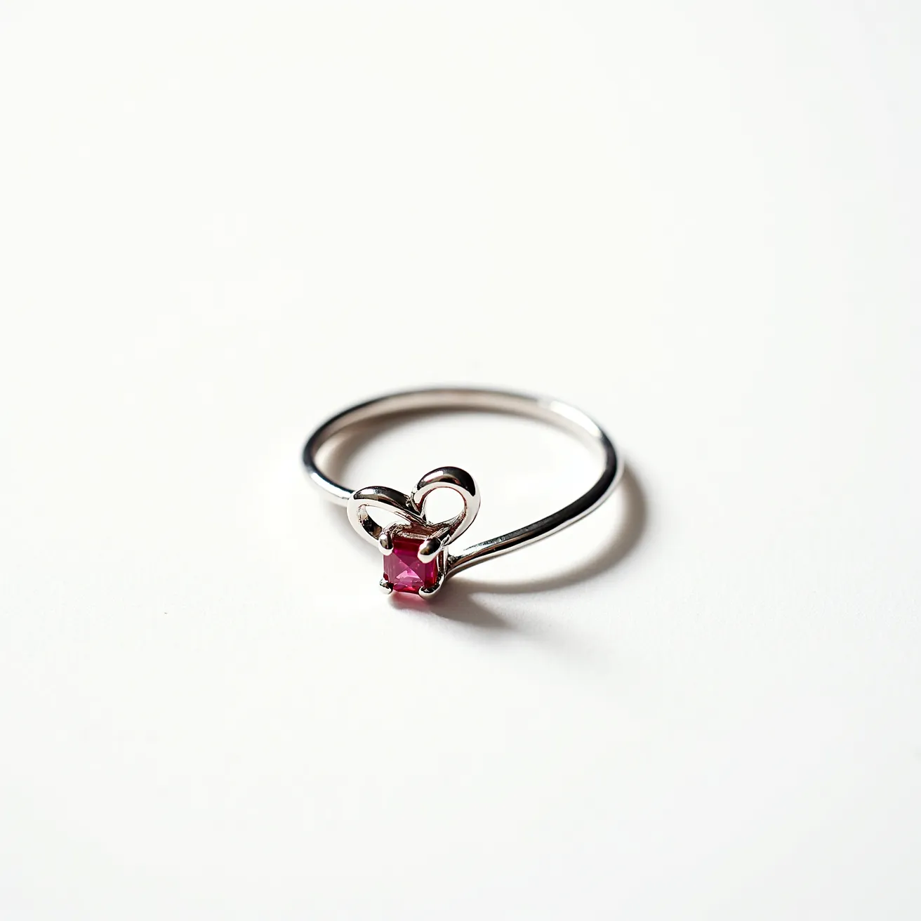 This heart wedding ring features a delicate band crafted from a silver-toned metal, elegantly shaped into a heart motif at the center. Set within the heart is a small, square-cut pink gemstone, likely a ruby, which adds a touch of vibrant color. The stone is held securely by a prong setting that enhances its visibility and brilliance. The design is simple yet elegant, with a focus on the heart and gemstone as the central decorative elements.