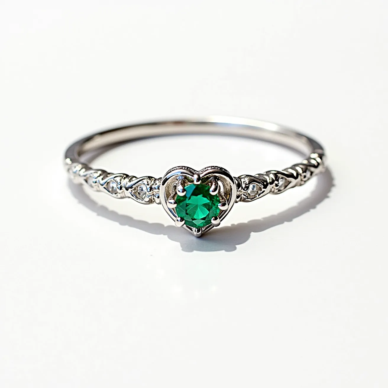 This heart wedding ring features a delicate band, likely made of white gold or platinum, adorned with small, round-cut diamonds set in a repeating heart pattern along the shank. At the center, a vibrant, round-cut green gemstone, possibly an emerald, is securely held in a six-prong setting encased within a heart-shaped design. The central stone's setting allows for maximum light reflection, enhancing its brilliance. This elegant design combines classic elements with a modern twist, making it a unique and meaningful piece.