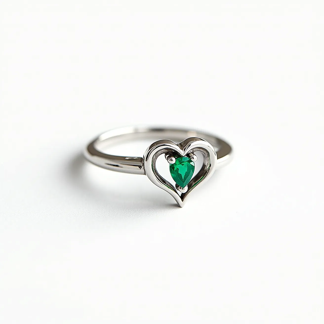 This heart wedding ring features a sleek metal band, possibly made of white gold or platinum, shaped into an elegant heart design. At the center of the heart lies a vibrant, green, pear-shaped gemstone, likely an emerald, securely set with a three-prong setting that enhances its brilliance. The combination of the metal and the stone creates a sophisticated and romantic look, making this ring an ideal symbol of love and commitment.