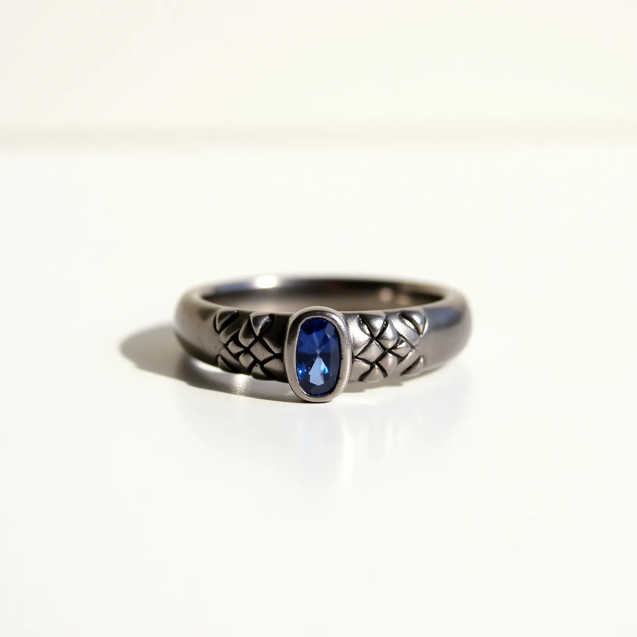 This hematite ring features a dark, metallic band with intricately engraved patterns along its surface. At its center, a striking oval-cut blue gemstone is set securely in a bezel setting, adding a vibrant contrast to the cool tone of the hematite. The smooth, polished finish of the band enhances the elegance of the design, complementing the gemstone's reflective qualities. The craftsmanship showcases a balance between modern and classic elements, making it a versatile piece suitable for various occasions.