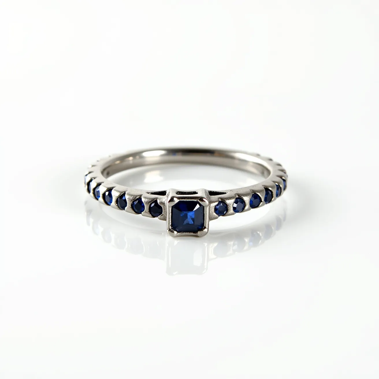 This hematite ring features a sleek and polished appearance, set with a series of small, dark blue gemstones that are uniformly placed around the band. The central stone is square-cut, held securely in a prong setting that enhances its visibility and brilliance. The surrounding stones are round-cut, similarly set to create a continuous, elegant design. The band's metallic finish complements the overall cohesion of the piece, emphasizing the stylish contrast between the hematite and the dark blue stones. There are no clasps or additional attachments, as the ring is designed in a classic circular form.