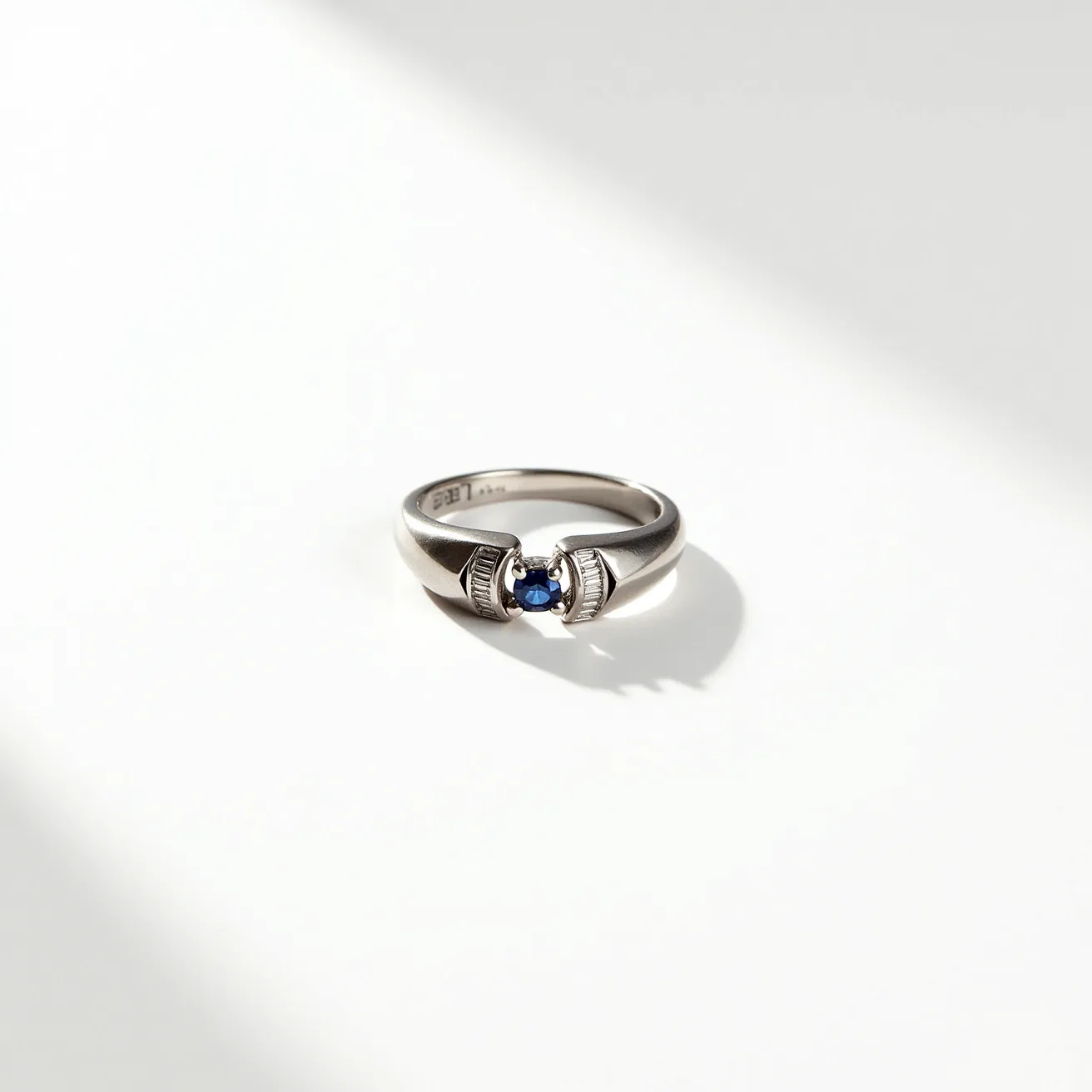 This hematite ring features a polished band and is accented with a central blue gemstone, likely a sapphire, set in a bezel setting that securely holds the stone in place. The round cut of the gemstone enhances its brilliance, and the ring’s sleek design complements the stone’s vibrant color. There are no additional clasps or attachments on the ring, allowing the focus to remain on the central gem and the smooth hematite surface.