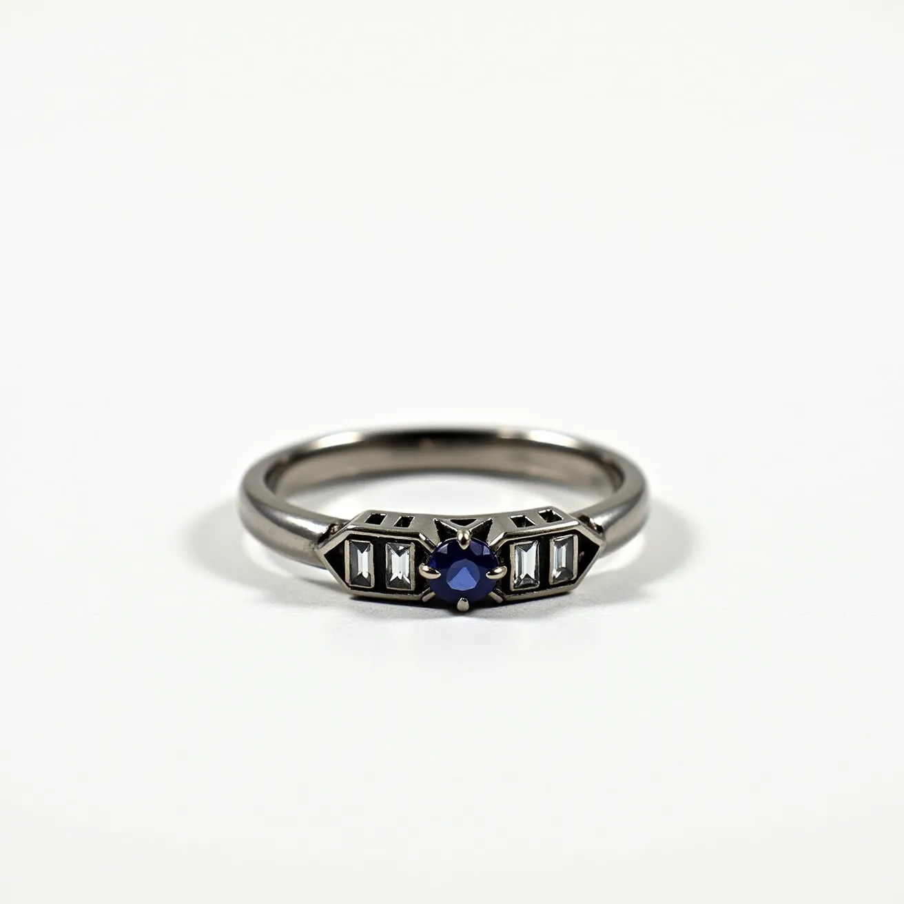 This hematite ring features a metallic band that supports an elegant arrangement of gemstones. At the center is a round-cut blue sapphire, held securely by prongs in a star-shaped setting. Flanking the central sapphire are rectangular baguette-cut clear stones set symmetrically, enhancing the ring's geometric appeal. The design is sleek and modern, with a polished finish that complements the contrasting colors of the stones and band, creating a striking visual effect without any additional clasps or attachments.