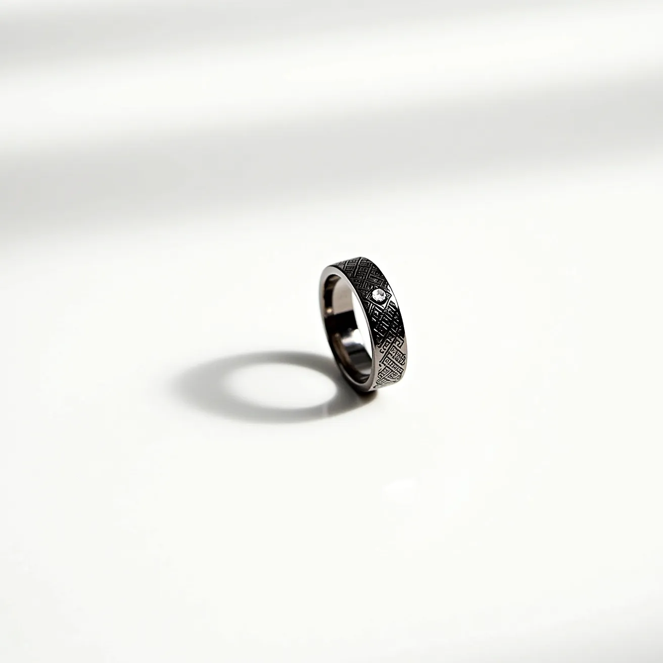 This hematite ring features a polished dark metallic surface typical of hematite, offering a sleek and modern appearance. The ring is adorned with a single small round-cut gem, set into the surface without any visible prongs, creating a smooth and integrated look. The band displays a geometric engraved pattern that adds texture and visual interest. The ring seems to be a simple slip-on style, with no clasps or additional attachments, emphasizing its elegant and understated design.