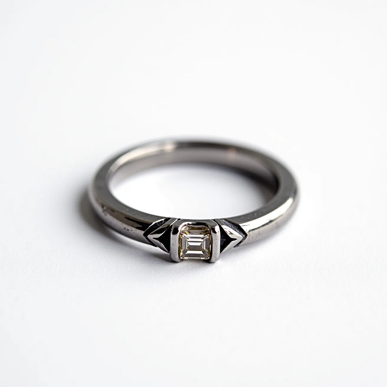 This hematite ring features a sleek metallic band with a polished finish, showcasing a minimalist yet elegant design. At its center, there is a single, rectangular gemstone that appears to be set in a bezel setting, providing a secure and smooth appearance. The gem is cut in an emerald shape, lending the ring a sophisticated and modern aesthetic. The hematite material gives the ring its distinctive gray sheen, enhancing its contemporary look. There are no additional clasps or attachments, highlighting the ring's streamlined and classic design.