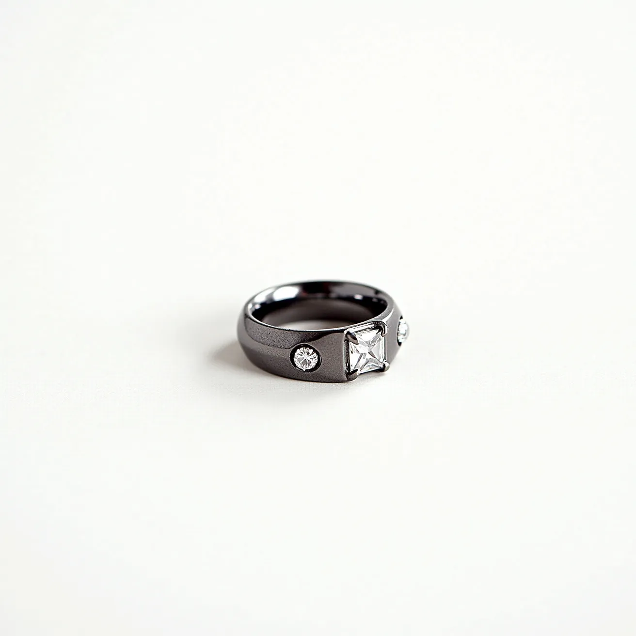 This hematite ring features a smooth and polished band, showcasing a distinctive metallic gray luster characteristic of hematite. The ring is adorned with a prominent square-cut gemstone positioned centrally, flanked by two smaller round-cut stones on either side. The square-cut gem is set in a sturdy bezel setting that securely holds the stone, while the round gems are accented in flush settings, giving a streamlined and minimalistic appearance. The combination of hematite with the clear gemstones creates a striking contrast, emphasizing the elegance and modern aesthetic of the design.