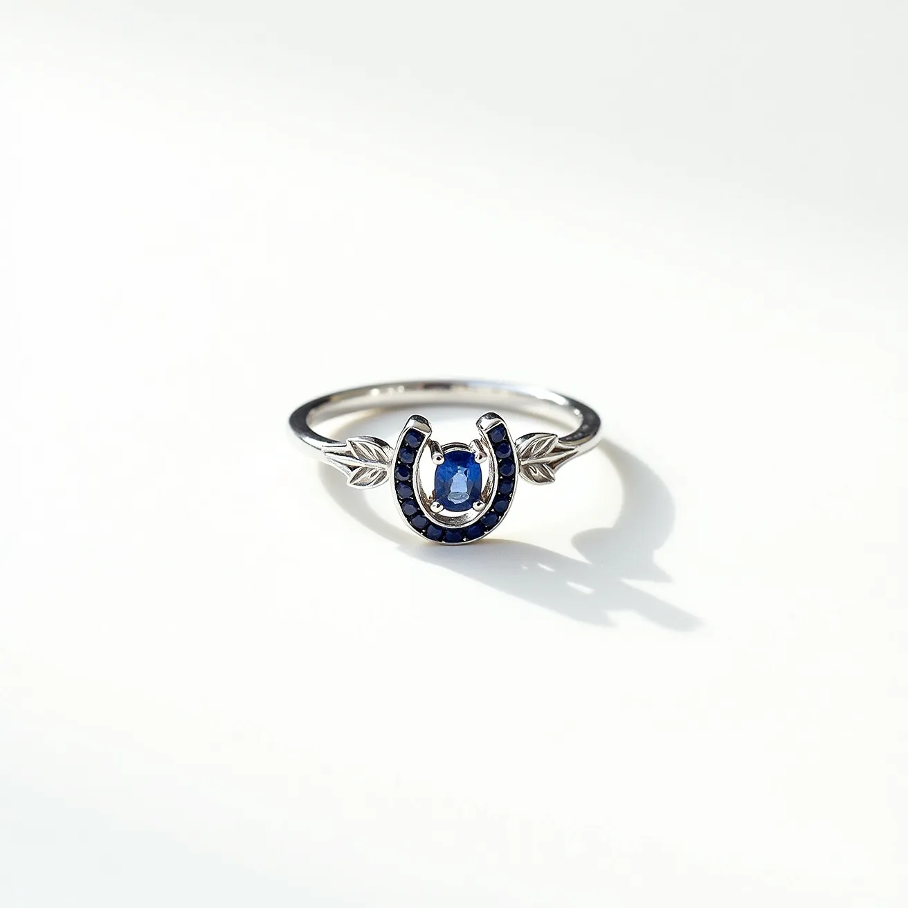 This horseshoe ring features a sleek metal band, likely made of sterling silver or white gold, that elegantly holds a horseshoe design at its center. The horseshoe is adorned with small, round-cut blue sapphires, which are bead-set, creating a vibrant yet understated sparkle. At the center of the horseshoe sits an oval-cut blue sapphire, prong-set to enhance its brilliance and centrality. Flanking the horseshoe design are two leaf-shaped accents, also crafted from the same metal as the band, which add an element of nature-inspired detail to the piece, giving it a distinctive and elegant look.