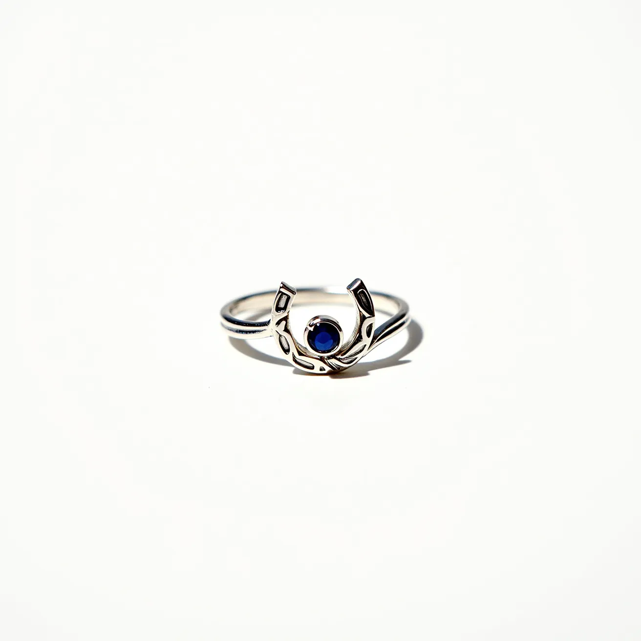 This horseshoe ring features a sleek metallic band crafted from what appears to be silver. At its center lies a deep blue gemstone, possibly a sapphire, which is round cut and securely set within the design. The horseshoe motif elegantly frames the gemstone, symbolizing luck and elegance. There's no visible attachment or clasp as it is designed as a traditional ring.