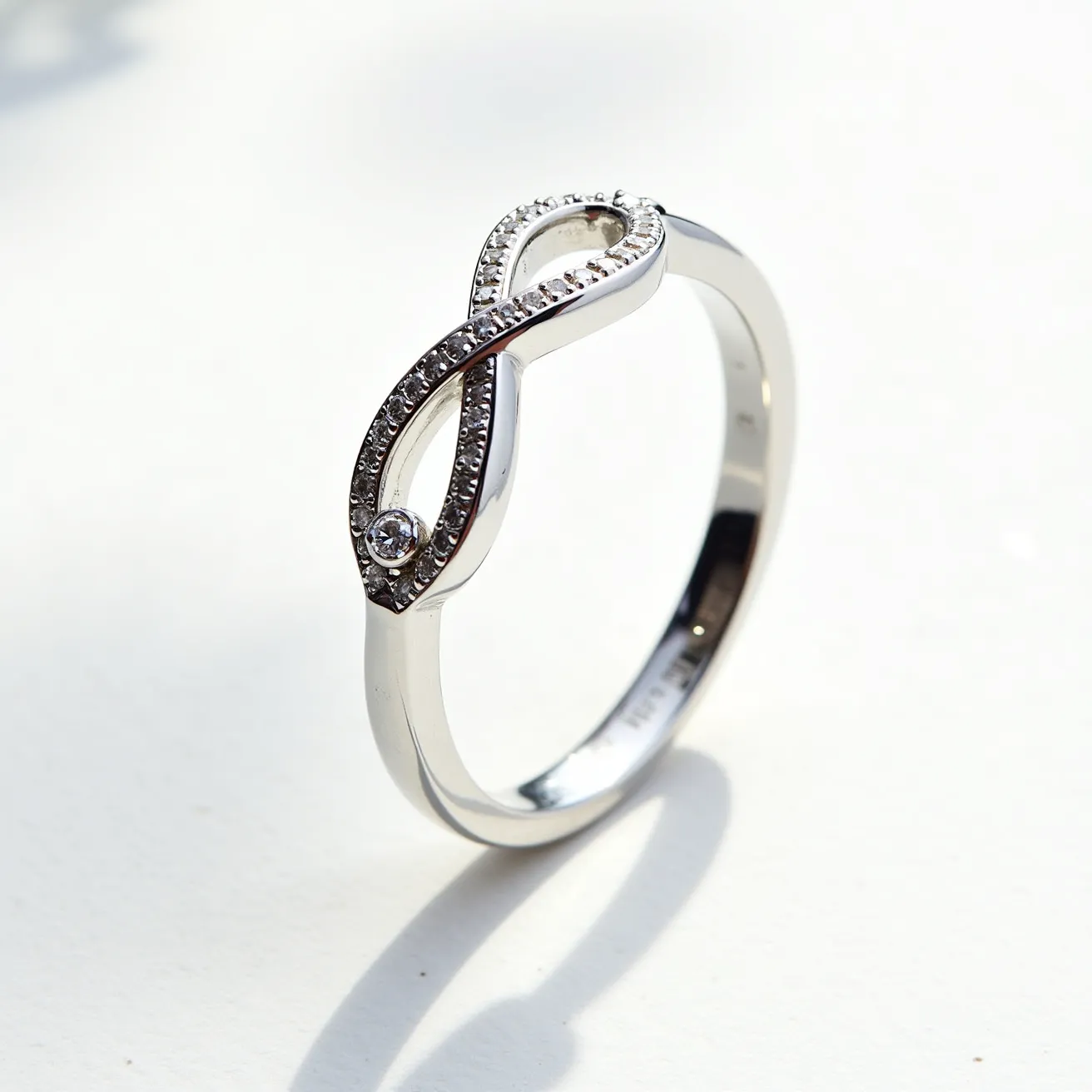 This infinity ring is crafted from a polished metal, possibly white gold or platinum, featuring an elegant infinity symbol as its centerpiece. The symbol is adorned with a row of small, round-cut diamonds set in a pavé style, enhancing its shimmering allure. A larger, singular round diamond is bezel-set at one end of the infinity loop, adding a striking focal point to the design. The ring band is sleek and smooth, complementing the intricate detailing of the infinity motif. The overall design presents a harmonious blend of simplicity and sophistication, perfect for special occasions or everyday elegance.