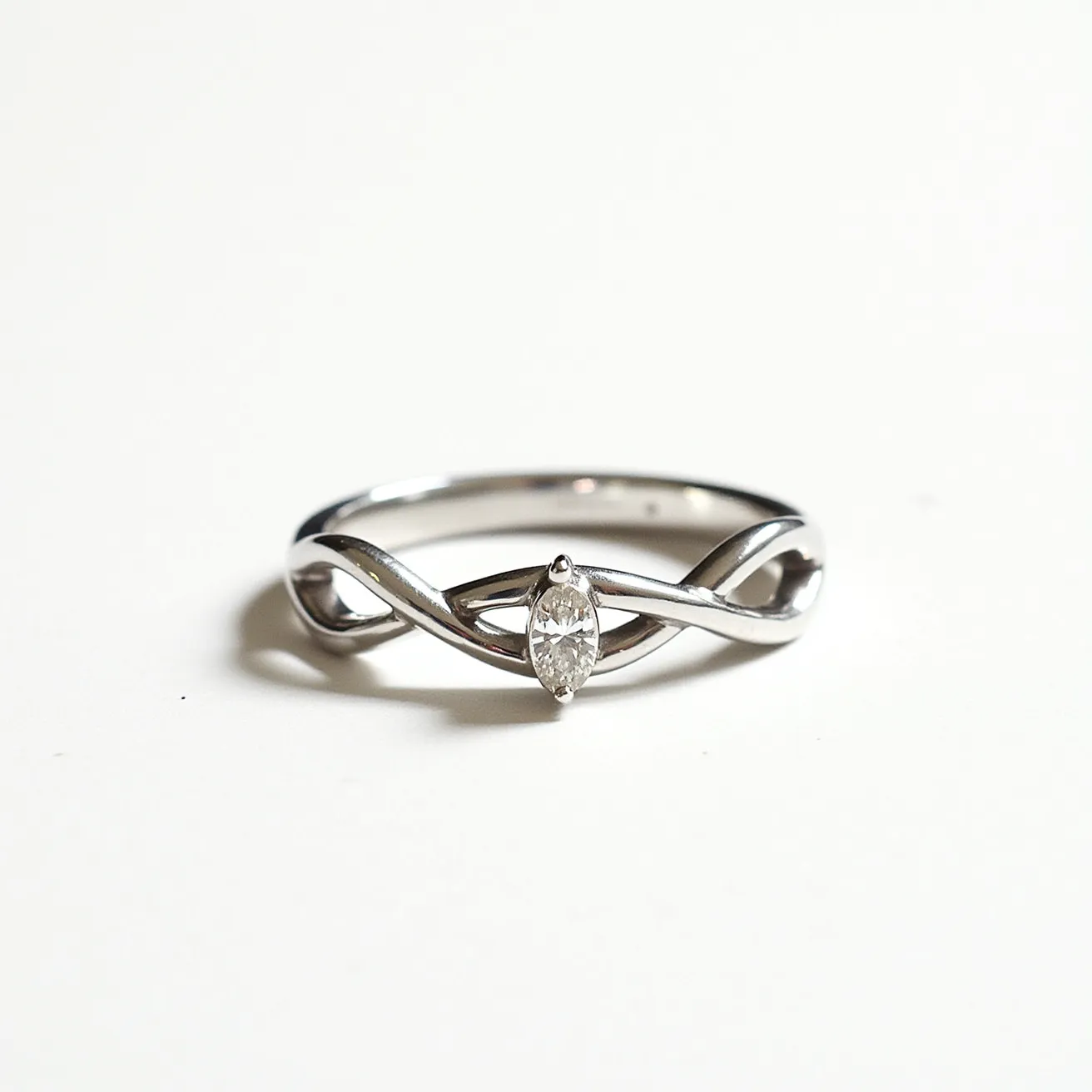 This infinity ring features a sleek, polished metal band, likely made of silver or white gold, crafted into an elegant infinity loop design. At the center of the infinity symbol, a marquise-cut diamond is set, reflecting a brilliant sparkle. The diamond is securely held in place by a prong setting, accentuating its elongated shape. The intertwining bands gracefully cradle the gemstone, creating a harmonious blend of geometry and luxury. The simplicity and sophistication of this design make it a timeless piece, ideal for symbolizing eternal love and commitment.