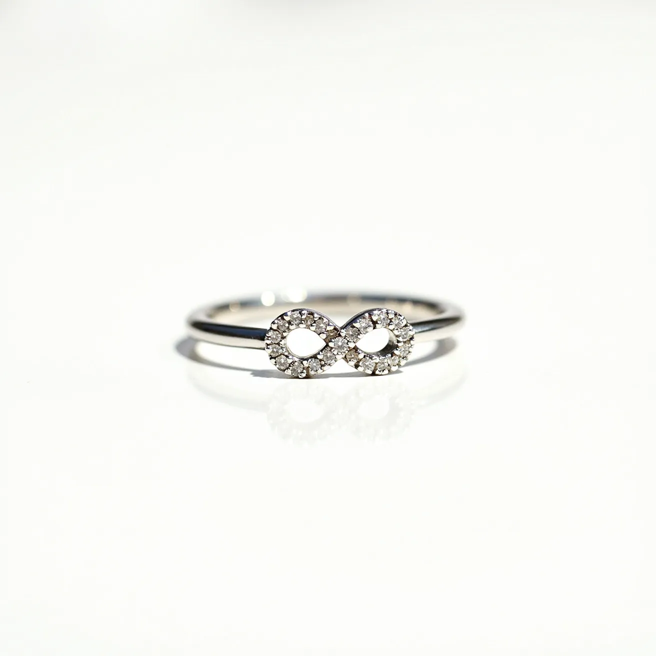 This infinity ring features a sleek metal band that is likely made of a polished white gold or sterling silver. The centerpiece is an infinity symbol encrusted with small, round, brilliant-cut gemstones that appear to be diamonds, set closely together in a pavé setting. There are no visible clasps or attachments, as the design is a continuous band signifying eternity. The arrangement of the stones provides a sparkling accent that contrasts elegantly with the smoothness of the metal band, making it an ideal piece to symbolize everlasting commitment or friendship.