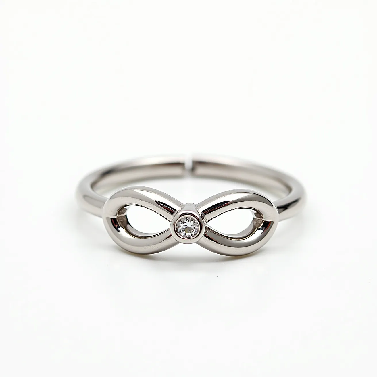 This infinity ring features a sleek design crafted from polished metal, likely stainless steel or white gold, forming the classic infinity symbol on the band. At the center of the infinity symbol, there is a small, round-cut gemstone set in a bezel setting, enhancing the elegance of the design with a minimalist touch. The ring does not appear to have a traditional clasp, as it is a continuous band designed to slip easily onto a finger. The reflective surface amplifies its sophisticated look, making it both a subtle and meaningful accessory.