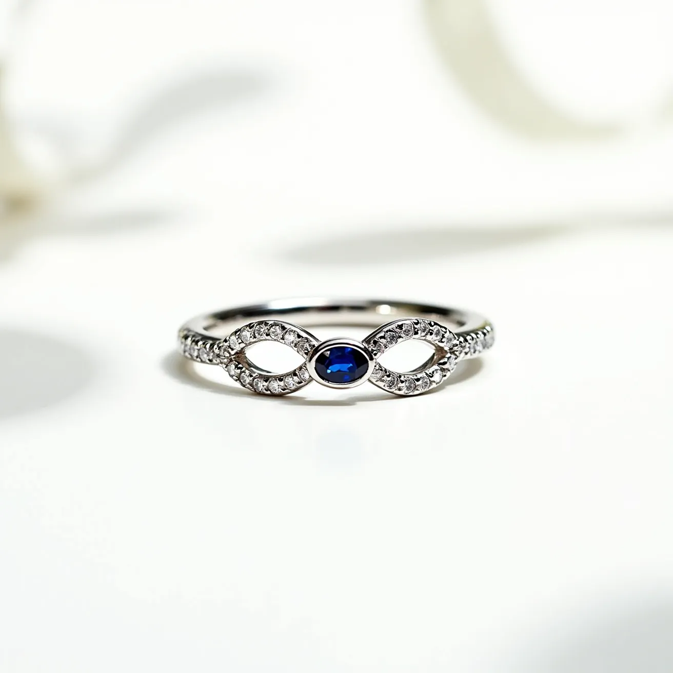 This infinity ring features a sleek band likely crafted from a polished metal such as white gold or platinum. It is designed in a continuous loop to symbolize eternity. The ring is adorned with a striking sapphire gemstone, cut in an oval shape and set in a bezel setting at the center of the infinity loop. Surrounding the sapphire, small round-cut diamonds are meticulously pavé set, enhancing the ring's elegance and brilliance by providing a sparkling contrast to the deep blue of the central gem.