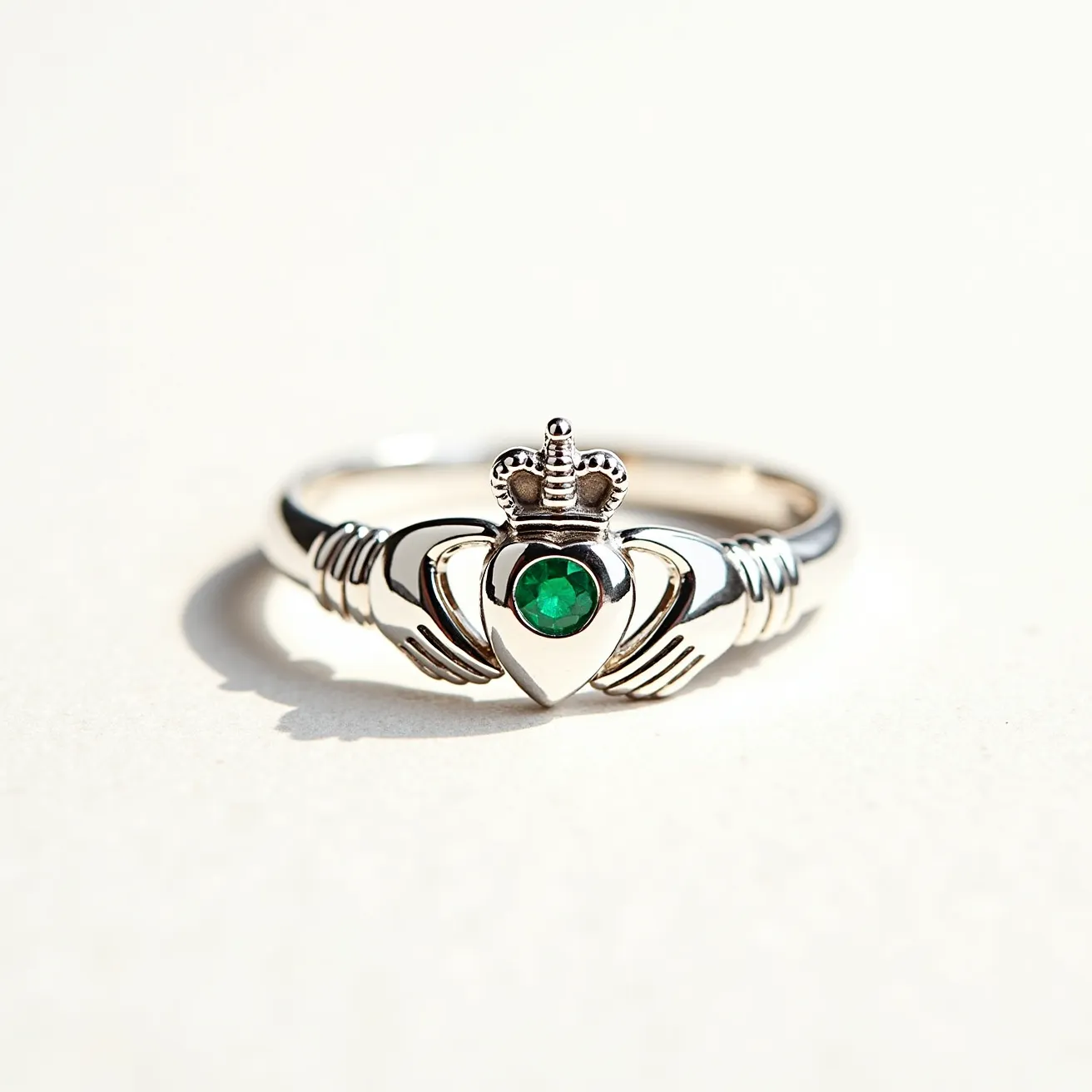 This Irish promise ring is crafted from silver and features a traditional Claddagh design with two hands holding a crowned heart. At the center of the heart is a round-cut green gemstone, likely an emerald, set in a bezel or prong setting, which adds a vibrant touch to the piece. The hands and crown are intricately detailed, emphasizing the symbolic elements of love, loyalty, and friendship inherent in this type of ring. The band itself is smooth and polished, providing a comfortable fit.