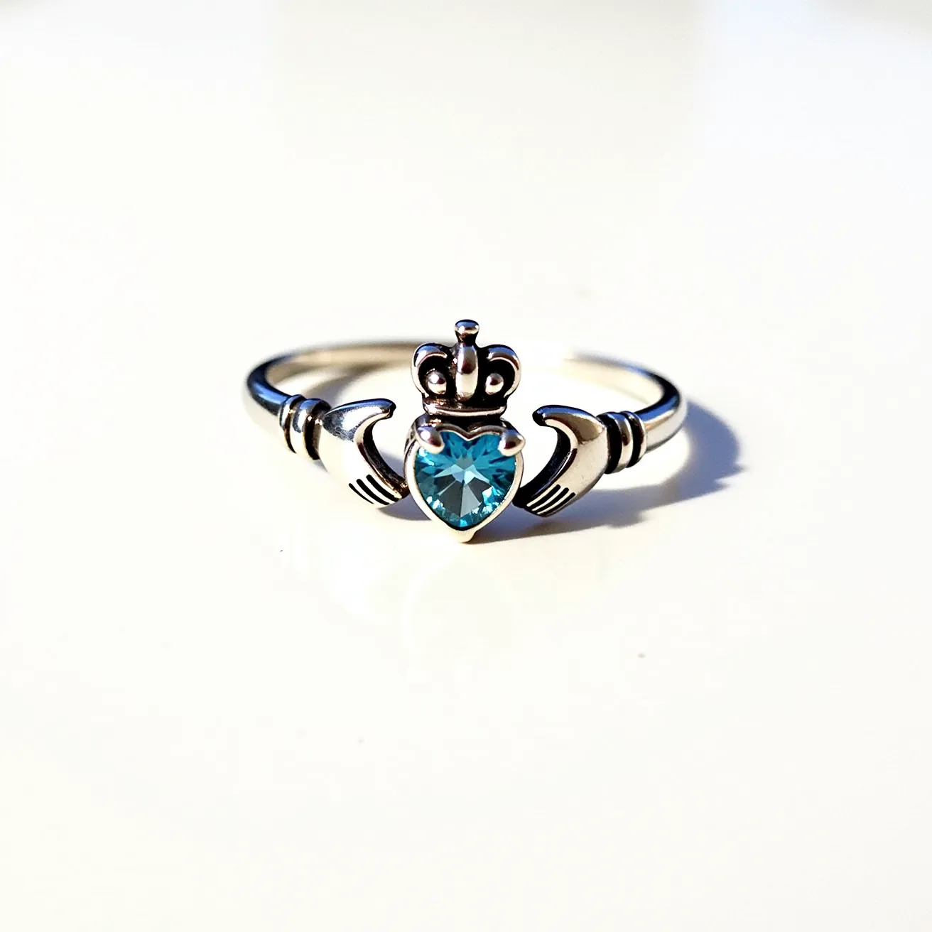 This Irish promise ring features a traditional Claddagh design, crafted from polished silver. At the center, a heart-shaped blue gemstone is set in a prong setting, symbolizing love. The heart is embraced by hands representing friendship, and topped with a crown denoting loyalty. The metalwork incorporates intricate detailing in the crown and hands, enhancing the elegance of this symbolic piece.