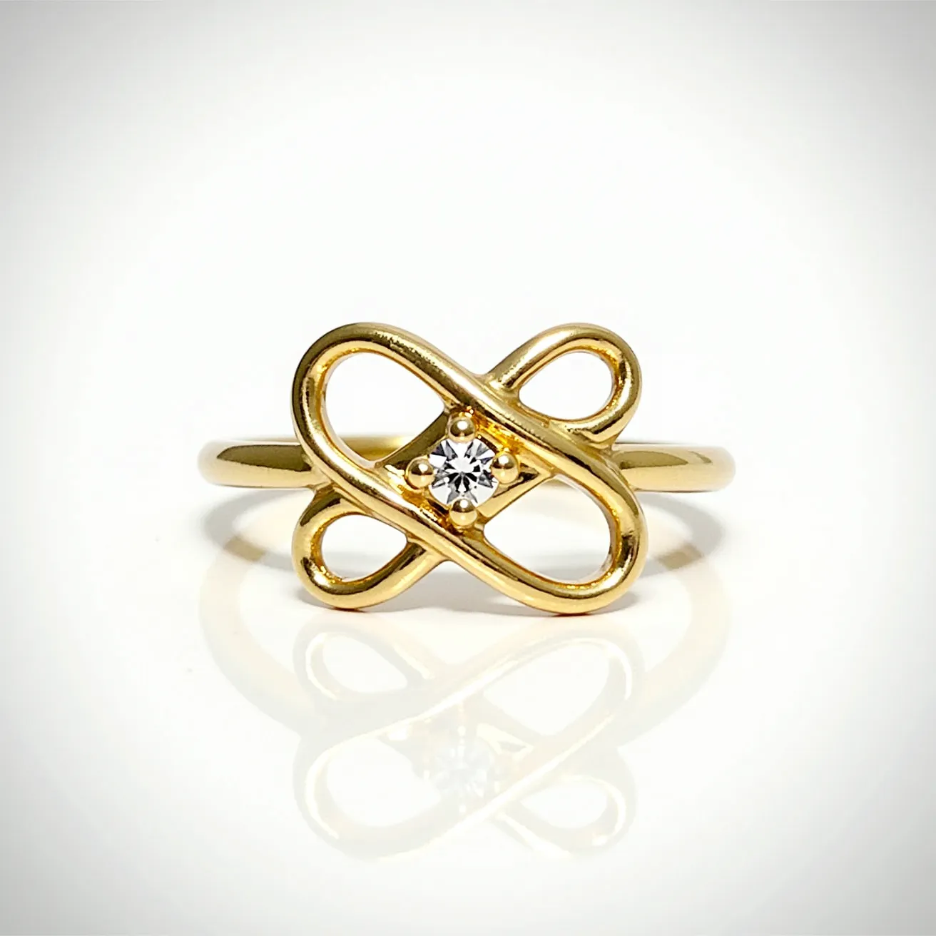 This knot ring is crafted from polished gold, featuring an elegant looped design that creates an intricate and symmetrical pattern. At its center, the ring proudly displays a small, round-cut diamond, securely held in place by a bezel setting that enhances its brilliance. The smooth band seamlessly integrates with the knot motif, providing a cohesive and stylish look without the need for additional clasps or attachments. The overall effect is both modern and timeless, with the diamond adding a touch of sparkle to the sophisticated gold structure.