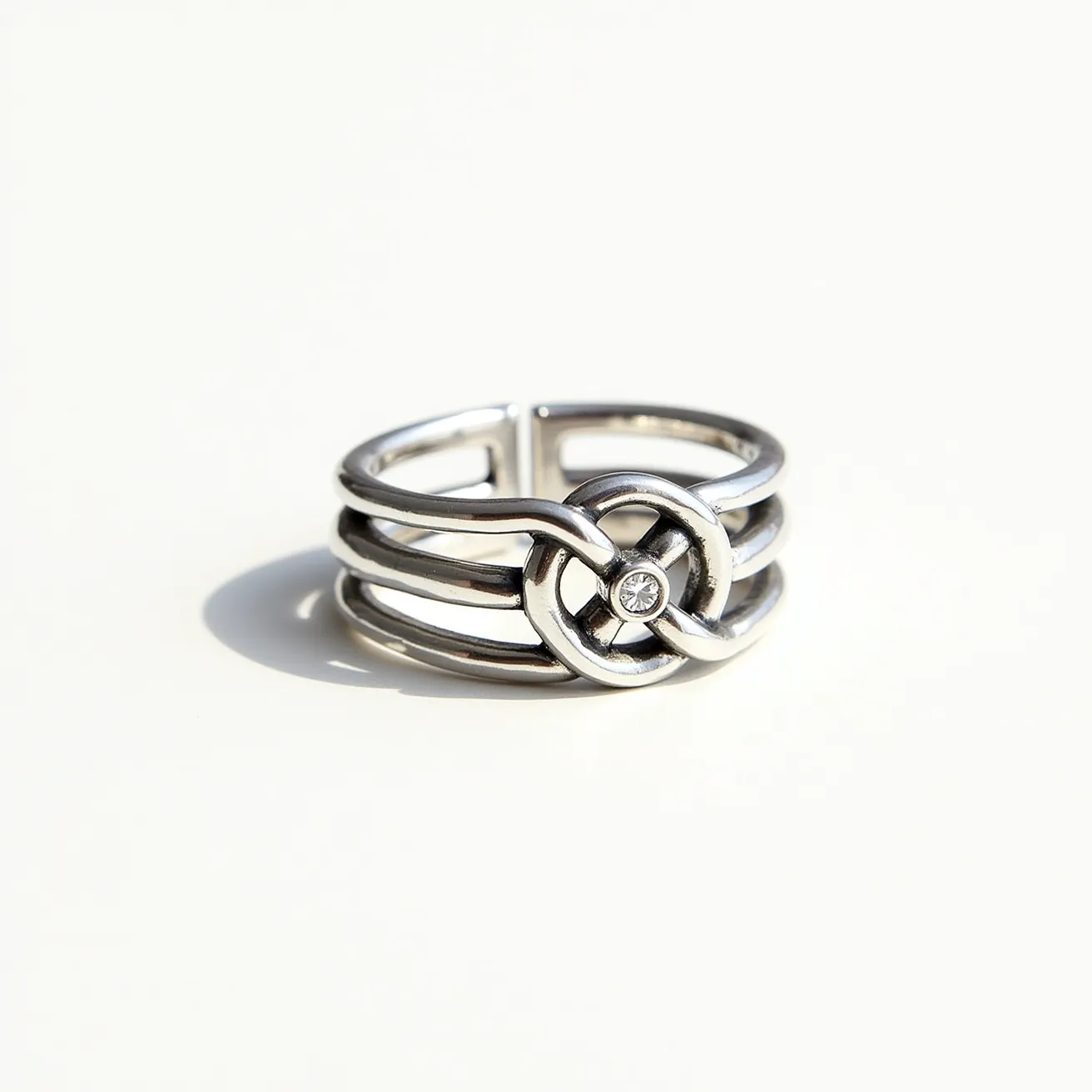 This knot ring features a sleek, modern design crafted from polished metal, likely silver or white gold, characterized by its smooth and reflective surface. The band splits into three strands, elegantly interwoven to form a knot at the center, adding a touch of sophistication and intricacy. At the heart of the knot is a small, round gemstone, possibly a diamond, set within a secure bezel setting that highlights its brilliance. The ring does not have an apparent clasp, suggesting it is a continuous circular band for easy wear.