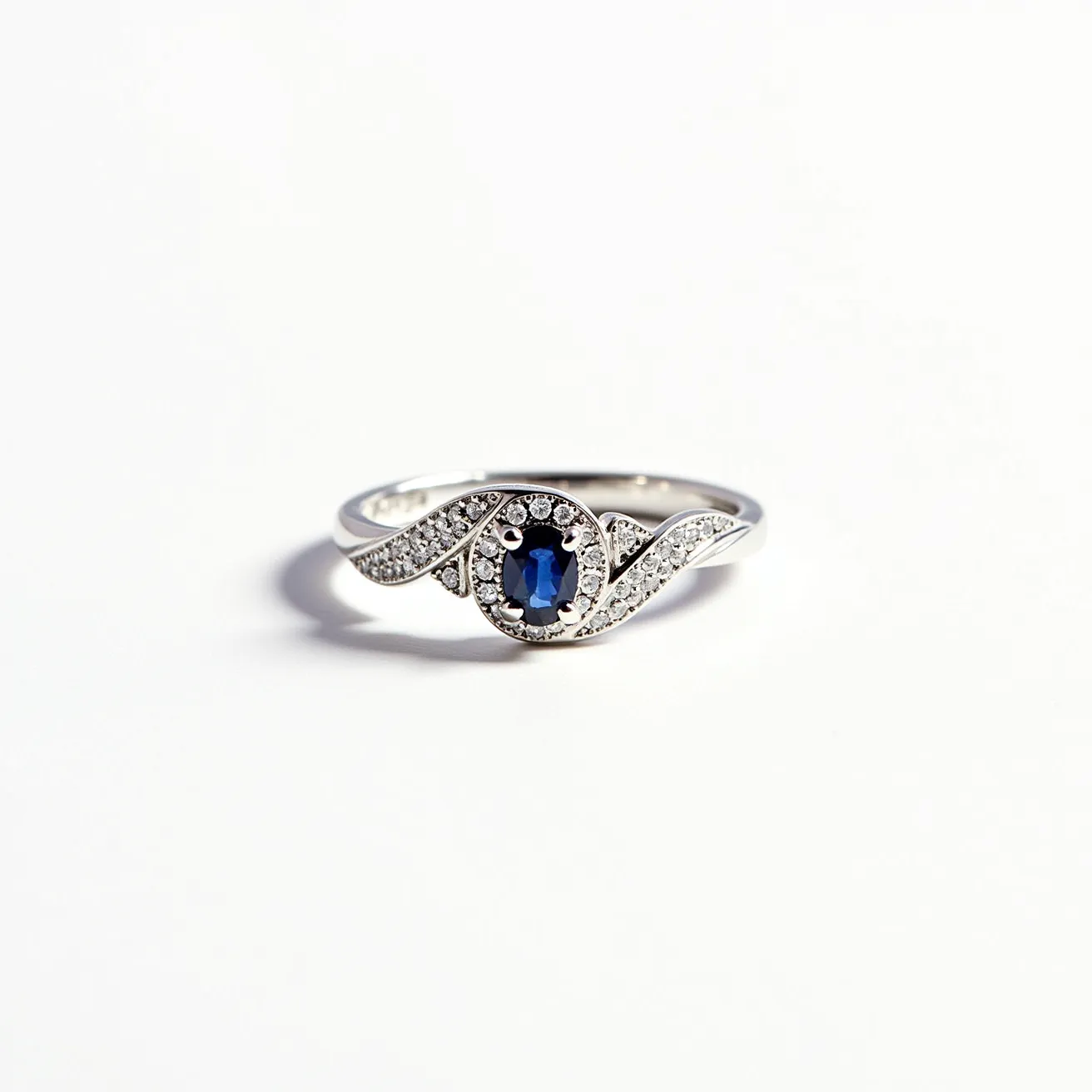 This love ring features a central blue sapphire, shaped in an oval cut, which is the focal point of the design. The sapphire is surrounded by a halo of small, round-cut white diamonds, enhancing its brilliance and adding a touch of elegance. The band is crafted from a polished silver metal, possibly white gold or platinum, which complements the sparkle of the gemstones. Alongside the central setting, the band gracefully splits and is adorned with additional small diamonds, set in a pave style that adds a continuous shimmer. The overall design of the ring highlights a modern and graceful aesthetic, perfect for expressing a timeless love.