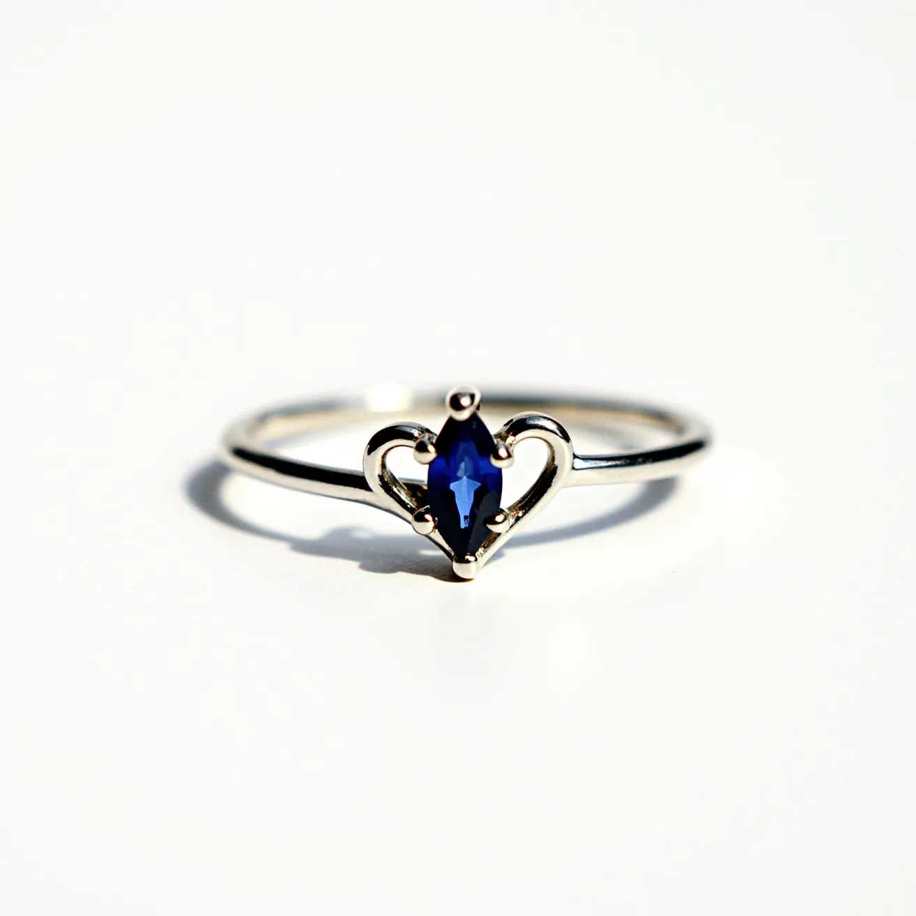 This love ring features a delicate band likely made of polished metal, possibly white gold or platinum, designed with an elegant simplicity. At its center, the ring showcases a marquise-cut blue gemstone, possibly a sapphire, securely held by a four-prong setting that accentuates its vibrant hue. Flanking the gemstone are gracefully curving elements that form a heart shape, further emphasizing its romantic theme. The craftsmanship of the ring combines elegance and sentimentality, making it a meaningful symbol of affection and commitment.