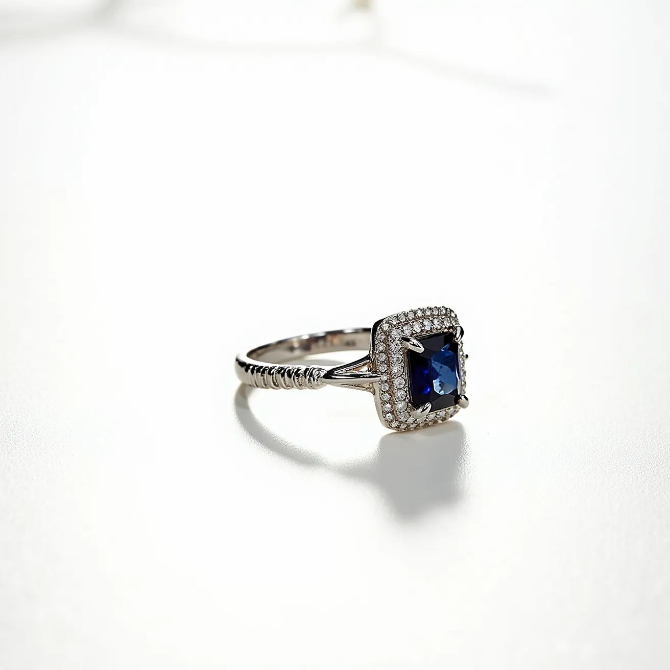 This March birthstone ring features a striking blue gemstone, likely aquamarine, cut into a square shape and set within a delicate prong setting. Surrounding the central gemstone is a halo of small, sparkling diamonds that enhance its brilliance. The band of the ring exhibits an intricate design, adding a touch of elegance to the piece. The ring is crafted from what appears to be white gold or platinum, complementing the cool tones of the stones. Overall, this piece combines elegance and sophistication, ideal for celebrating a March birthday.