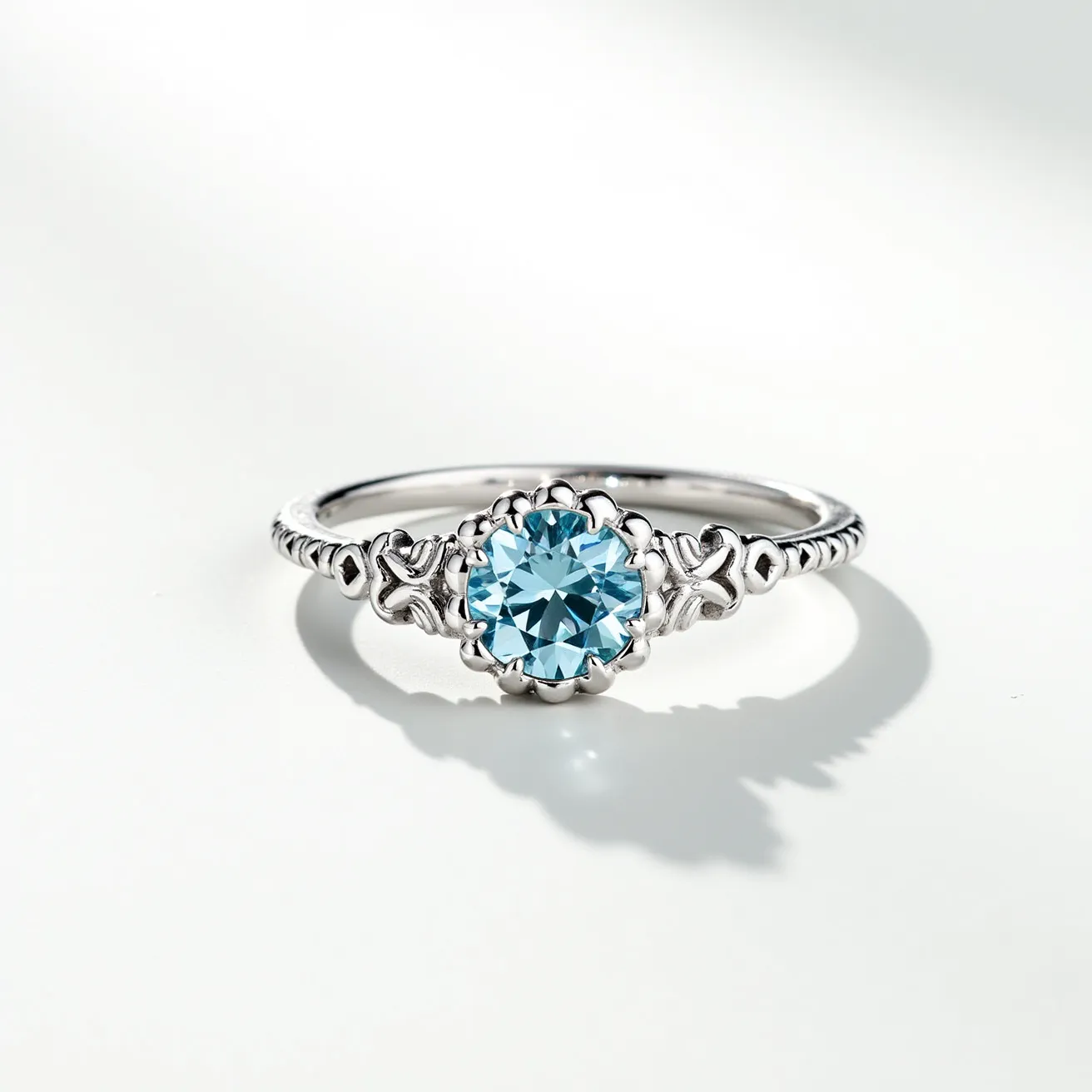 This March birthstone ring features a light blue aquamarine gemstone, expertly cut into a brilliant round shape. The stone is elegantly set into a delicate silver band, which incorporates intricate detailing along its sides. The gemstone is held securely in place by a floral-inspired setting, complementing the overall design with a touch of elegance. The band showcases an ornate design with a series of small embellishments that add to the ring's aesthetic appeal. This combination of aquamarine and silver creates a refined and sophisticated accessory, ideal for those born in March.