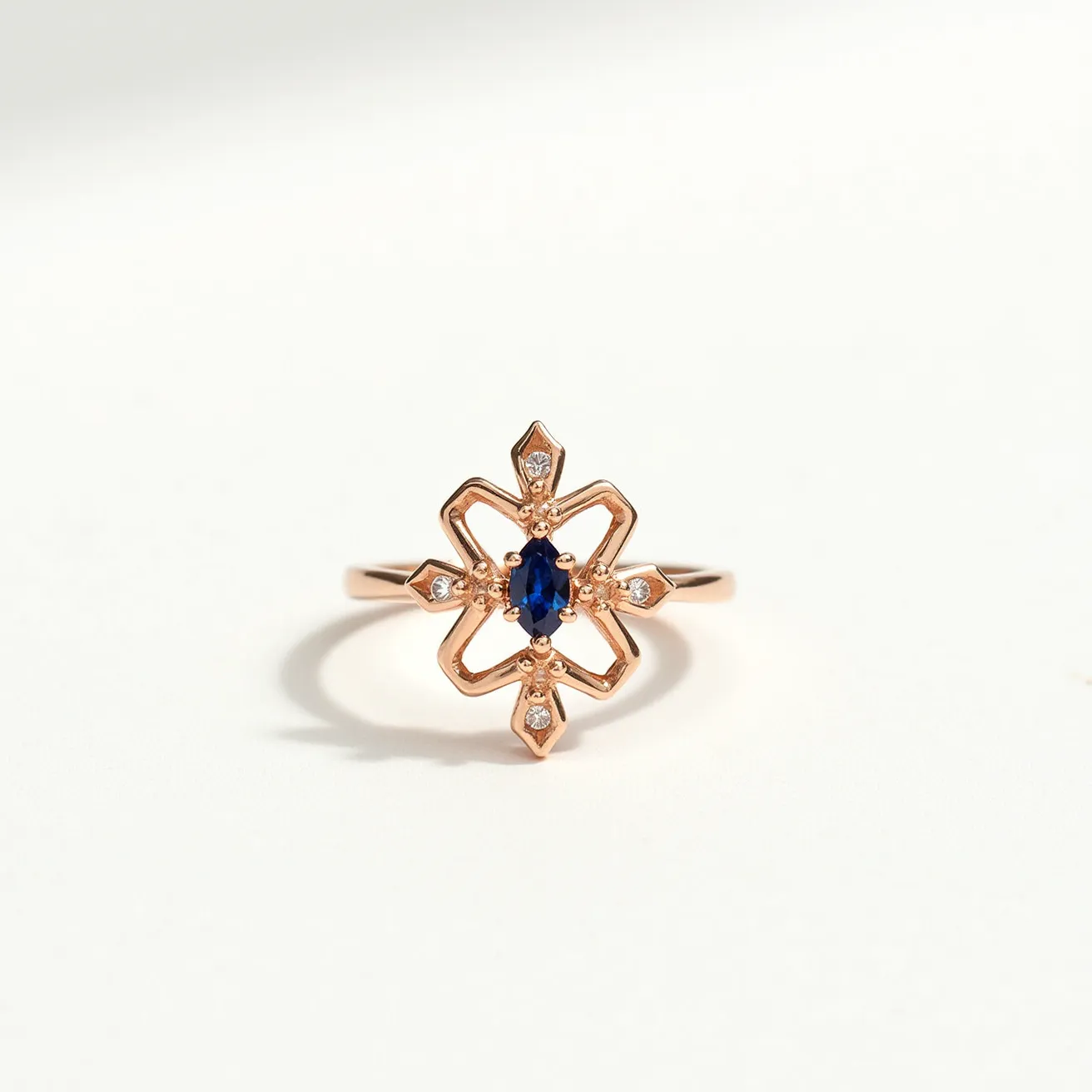 This marquise diamond ring features a central marquise-cut sapphire set in a geometric floral motif. The ring is crafted from rose gold, providing a warm, elegant backdrop for the central stone. Surrounding the sapphire are small round diamonds, each set at the tips of the star-like design, adding a touch of brilliance and contrast. The intricate openwork design complements the marquise cut of the sapphire, enhancing its elongated shape. The setting showcases a balance between the deep hue of the sapphire and the glittering diamonds, all held securely in a bezel setting with intricate metalwork, without any visible clasps or attachments.