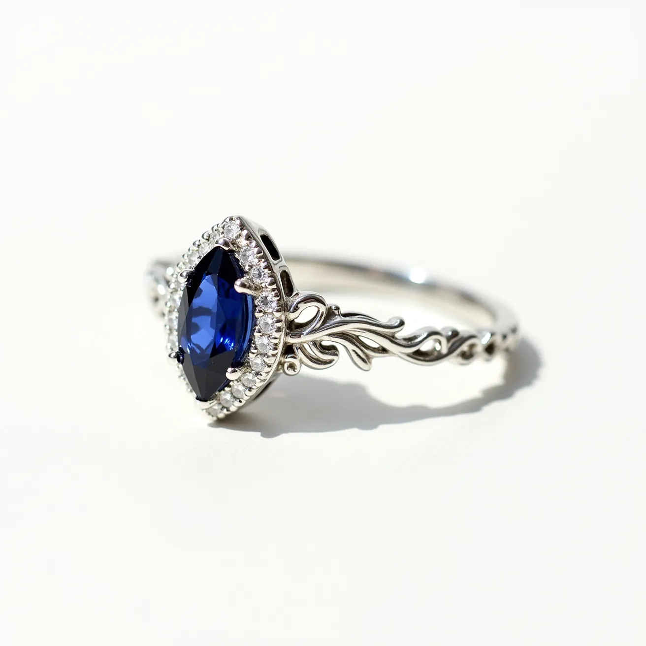 This marquise engagement ring features a striking marquise-cut blue gemstone as its centerpiece, elegantly surrounded by a halo of smaller round white diamonds. The setting and band are crafted from a polished silver-toned metal, with intricate detailing resembling delicate, flowing patterns on the sides. The metalwork complements the vividness of the center stone, enhancing its brilliance and appeal. The halo setting securely holds the blue gemstone in place, adding both elegance and stability to the design.