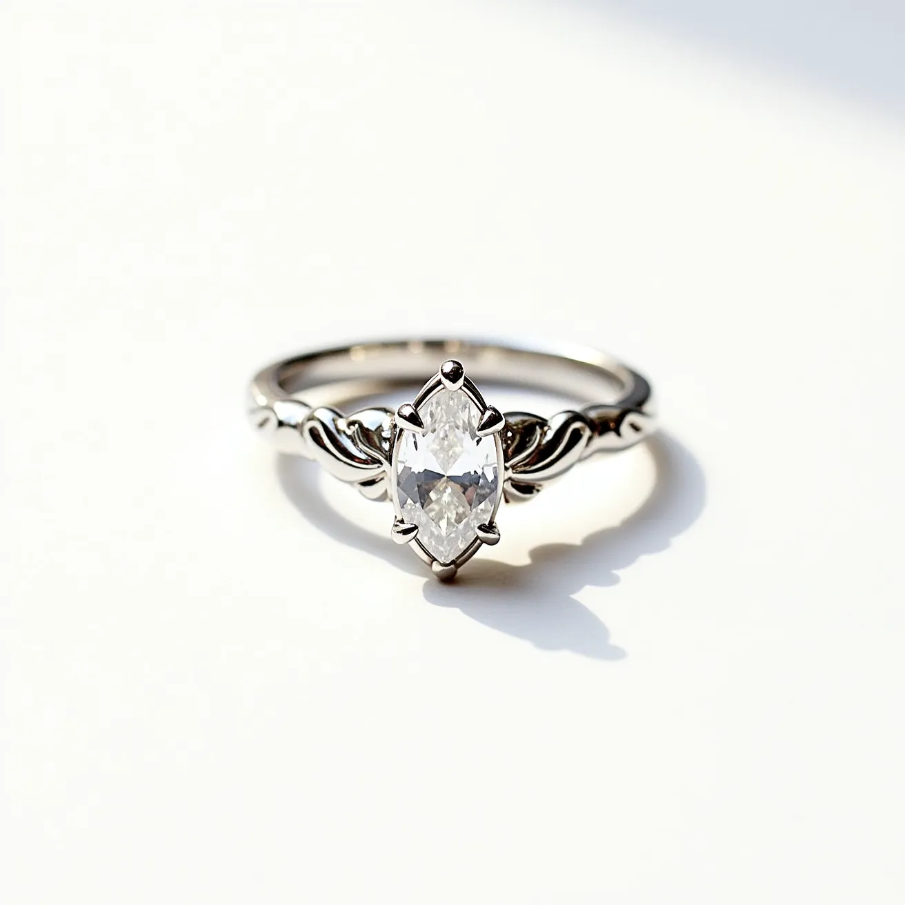 This marquise engagement ring features a central marquise-cut diamond, set in an elegant solitaire style. The stone is secured by prongs that accentuate the diamond's elongated shape, allowing for maximum light reflection. The band is crafted from a polished metal, possibly white gold or platinum, which complements the brilliance of the diamond. The band also incorporates subtle, decorative elements that enhance the ring’s overall aesthetic, lending it a touch of sophistication while maintaining focus on the striking marquise stone.