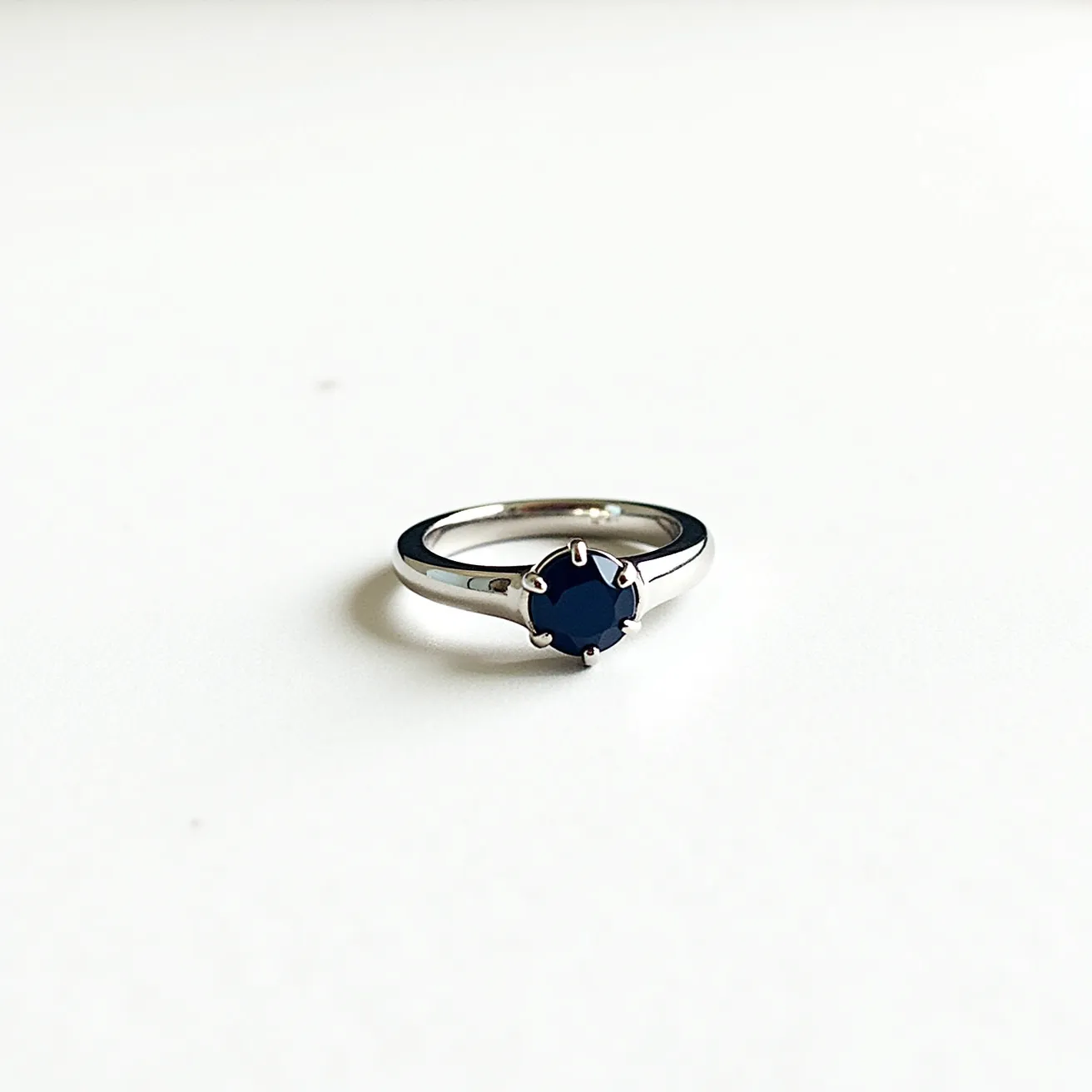 This men's engagement ring features a sleek, polished metal band, likely made of platinum or white gold. The band is elegantly simple, drawing attention to the striking centerpiece—a dark blue gemstone. The stone, which appears to be a round-cut sapphire, is secured in a classic six-prong solitaire setting, adding sophistication and prominence to the design. The prongs hold the gemstone firmly in place, ensuring its stability while maximizing its visibility and sparkle. This combination of materials and design creates a timeless piece that balances modern elegance with traditional style.