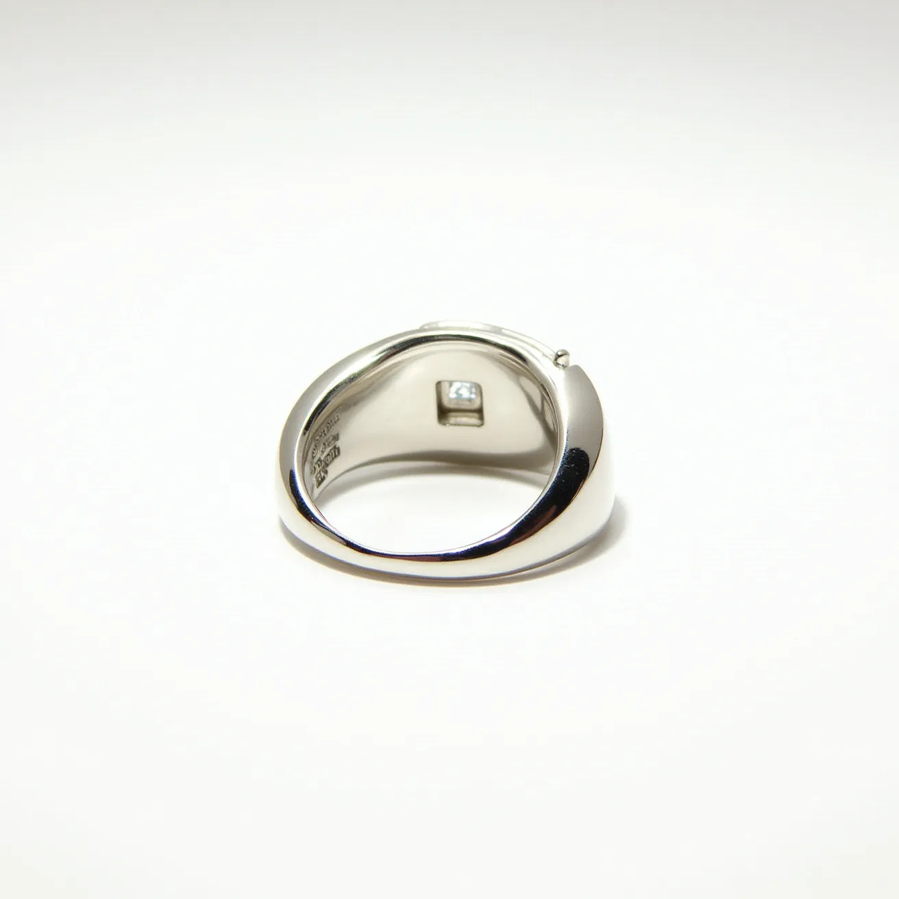 This men's pinky ring is crafted from polished metal, likely platinum or white gold, giving it a sleek and shiny appearance. Set subtly into the underside of the band is a small, square-cut diamond, which adds a discreet touch of luxury to the piece. The design is simple yet elegant, with a smooth finish and a slightly tapered band that enhances comfort and wearability. The ring does not feature any visible clasps or attachments, relying on its solid construction for security.