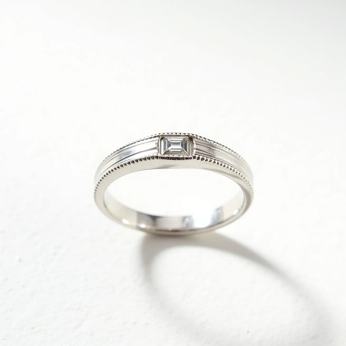 This men's pinky ring is crafted from a polished metal, likely silver or white gold, featuring a sleek band with intricate milgrain detailing along the edges. At its center, the ring showcases a rectangular baguette-cut clear gemstone, possibly a diamond, which is elegantly set in a bezel setting. The design combines modern simplicity with classic elegance, making it a subtle yet sophisticated accessory.