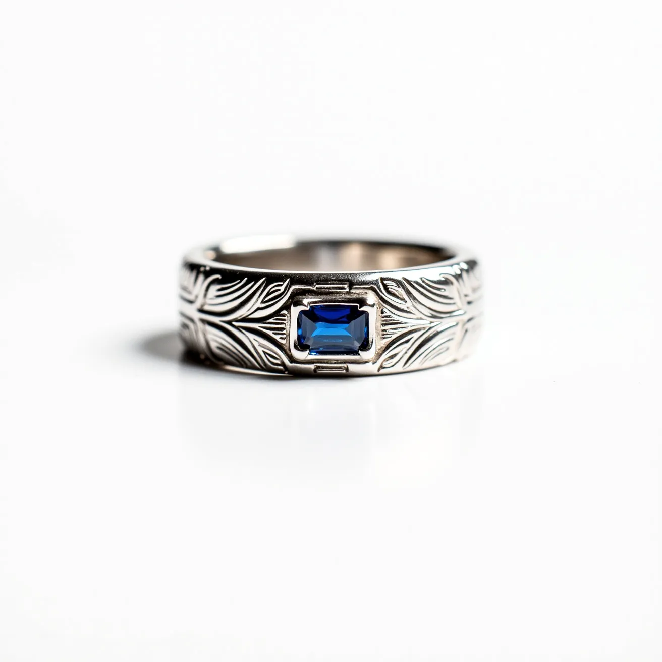 This men's ring features a design crafted from a metallic material, possibly silver or white gold, characterized by intricate engraved patterns that evoke a sense of elegance and sophistication. At its center, the ring showcases a rectangular, deep blue gemstone, likely a sapphire, which has been precision-cut to enhance its brilliance and sparkle. The gemstone is securely set within a bezel setting, which provides both functionality and style by encasing the gem in metal, ensuring its stability and protection. The band conveys a sense of refined artistry and classic masculinity through its meticulous detailing and the vibrant contrast provided by the centrally placed gemstone.