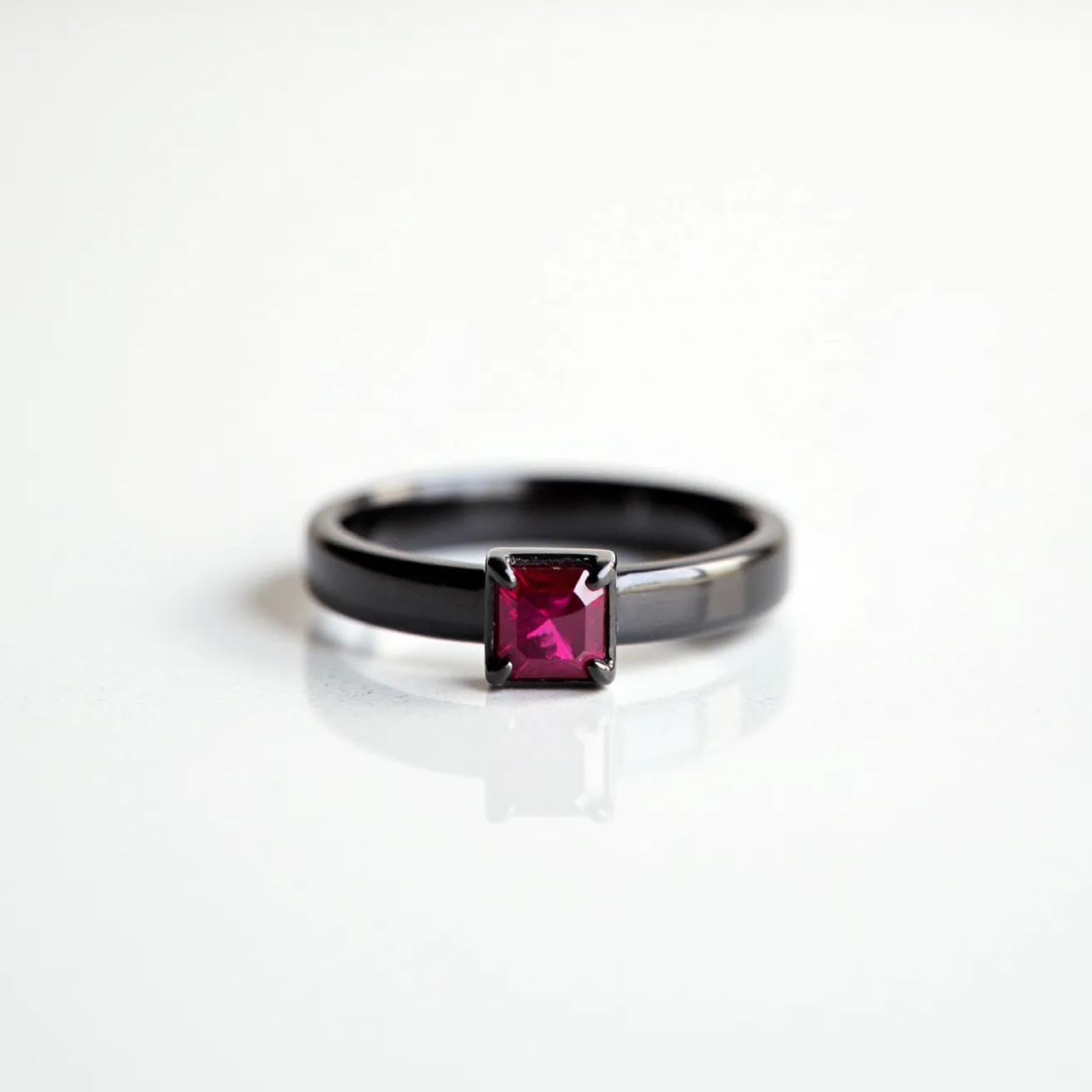 This men's ring features a sleek black band with a prominent square-cut red gemstone set at its center. The stone is held securely by a prong setting, enhancing its striking and vibrant appearance. The black band may be made of a modern material like tungsten or blackened steel, giving it a contemporary and sophisticated look. The simplicity of the band juxtaposed with the bold pop of color from the gemstone creates a balanced and elegant design, making it a standout piece for any occasion.