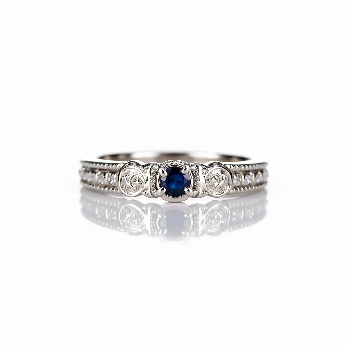 This men's ring features a white metal band, likely silver or white gold, adorned with intricate detailing along the edges. At the center, a round-cut blue sapphire is secured in a bezel setting, which provides both security and style. Flanking the central stone are smaller ornate carvings that add to the ring's classic and elegant design. The band displays a sequence of small, clear stones set in a row, possibly diamonds or cubic zirconia, enhancing the ring’s decorative appeal without any clasp or additional attachments.