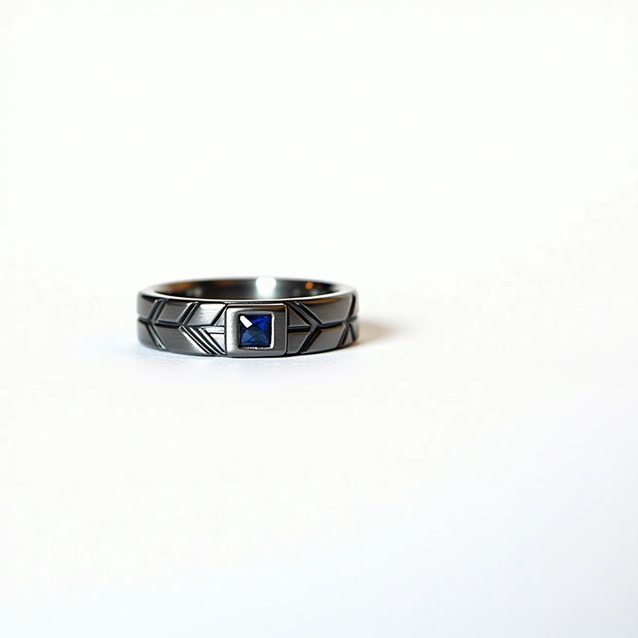 This men's ring exhibits a sleek metallic band adorned with geometric engravings, emphasizing its modern design. At the center lies a square-cut blue gemstone, likely a sapphire, intricately set in a bezel setting that securely frames the stone, allowing for both durability and elegance. The ring's combination of a polished metal finish and the vibrant blue of the gemstone creates a striking contrast, highlighting the gemstone's deep hue. The simplicity of its design with the geometric detailing adds an element of sophistication, making it suitable for both casual and formal attire.