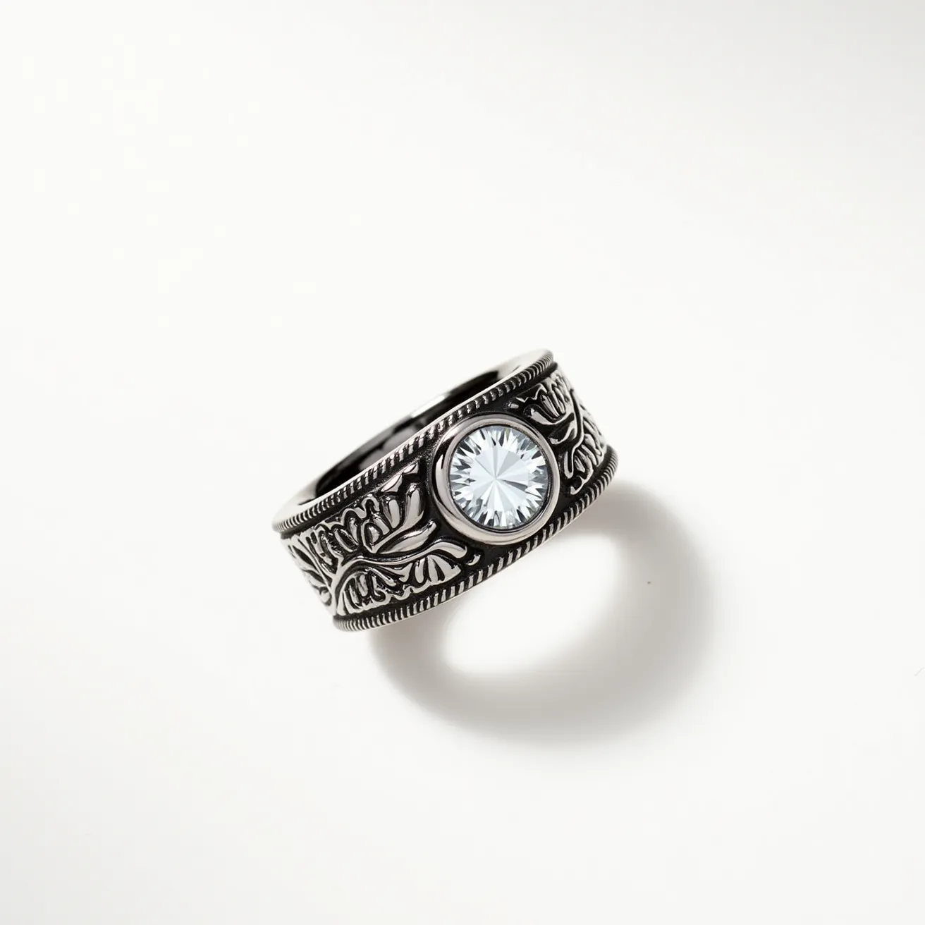 This men's ring features an intricate design with a dark metal band adorned with detailed engravings that resemble floral or ornate geometric patterns. At the center of the band is a prominent, circular-cut gemstone set in a bezel setting, which securely encases the stone. The gemstone, likely a clear crystal or diamond, serves as the focal point, reflecting light with its precise facets. The overall craftsmanship of the ring conveys a sense of elegance and strength, combining both decorative artistry and durability in its design.