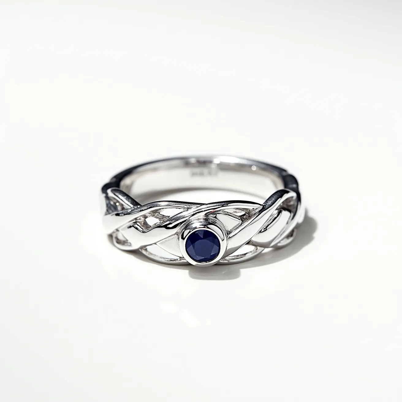 This men's ring features an intricate, braided design crafted from polished silver, showcasing a modern and elegant appearance. Centrally set within the ring is a deep blue gemstone, likely a sapphire, which is cut in a round shape and secured with a bezel setting that provides a sleek and secure display. The ring exhibits a seamless and continuous band, emphasizing its unity and craftsmanship without any visible clasps or additional attachments.